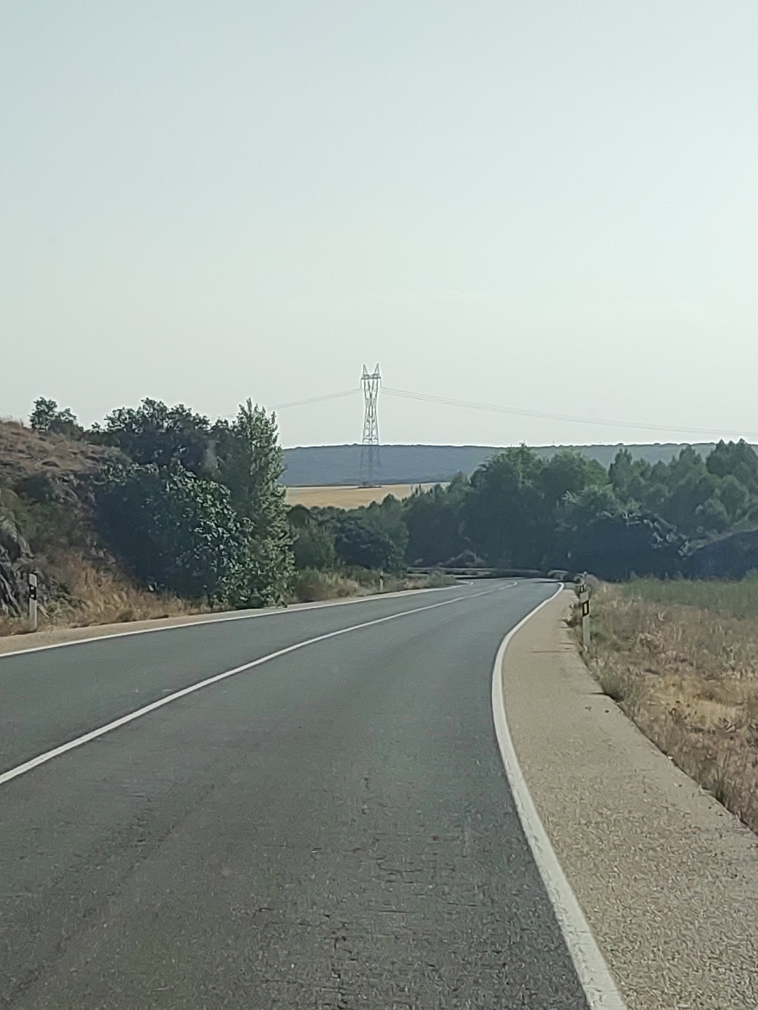 The image shows a paved road curving to the right, with a white line marking the lane. The road is bordered by a sandy shoulder on the right and a rocky, overgrown embankment on the left. In the distance, a tall pylon supports power lines. The landscape is dry and appears to be rural, with sparse vegetation, including bushes and trees. The sky is pale and hazy, suggesting a bright, possibly warm day. There are no people or vehicles visible. The scene is likely a photograph taken from a moving vehicle, given the perspective and slight motion blur. The location is identified as being in the city of Saelices, Spain.