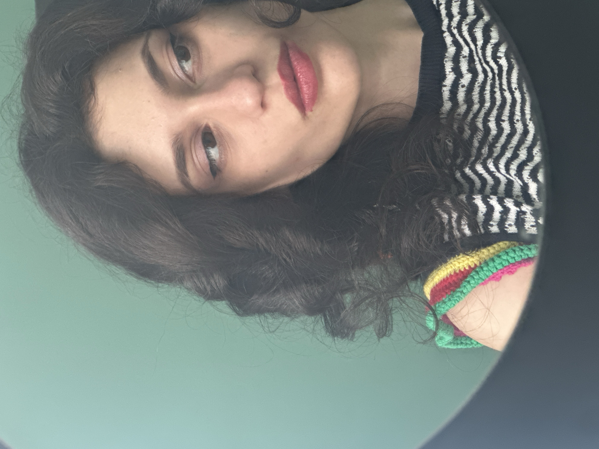 The image shows a close-up, waist-up shot of a young woman with dark, curly hair and light skin. She is wearing a black and white striped top with a crocheted, multi-colored trim on the shoulder, featuring bands of green, yellow, red, and pink. Her makeup includes eyeliner and pink lipstick. The background is a plain, light green wall. The woman is reflected in what appears to be a dark, circular frame, possibly a mirror or a ring light. The scene is indoors, with no visible city elements.