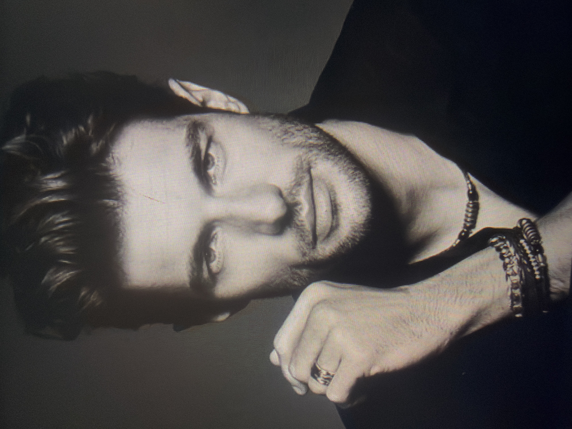 A black and white photograph shows a man with stubble, gazing slightly downward. His head rests on his hand, adorned with a ring. He wears beaded bracelets and a necklace. The location is unspecified and there is no indication of it being Sofia, Bulgaria.