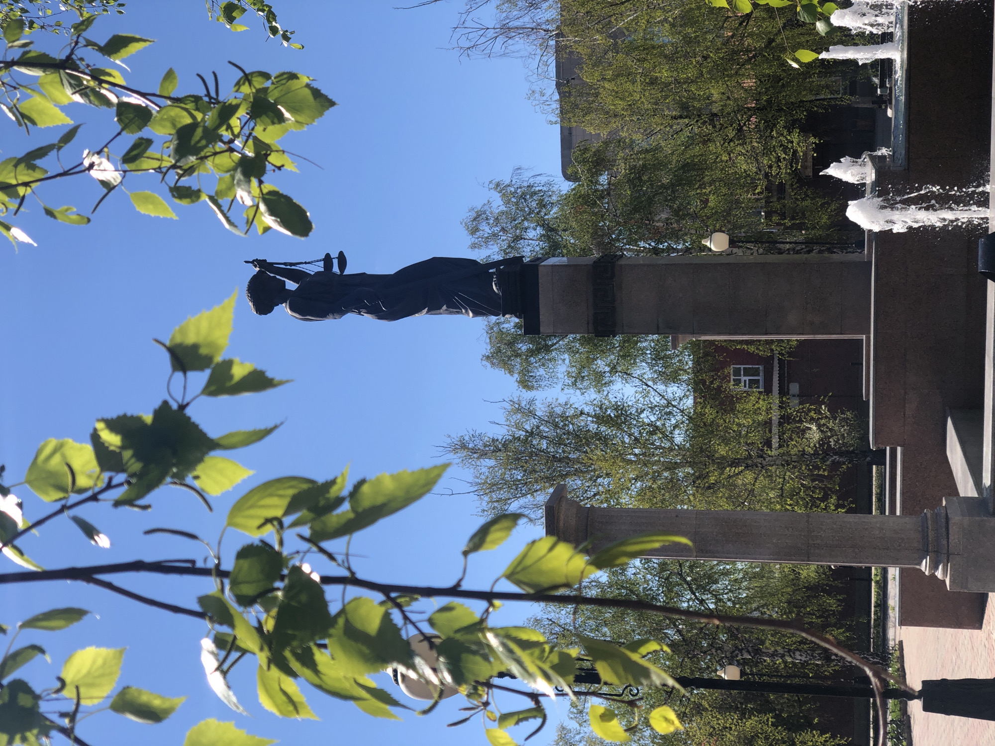 The image captures a view of the statue of Lady Justice in Krasnoyarsk, Russia. The statue, atop a column, holds scales. The statue appears to be made of dark metal. The background features trees, buildings, a fountain with water jets, and a clear blue sky. A branch with bright green leaves is in the foreground.
