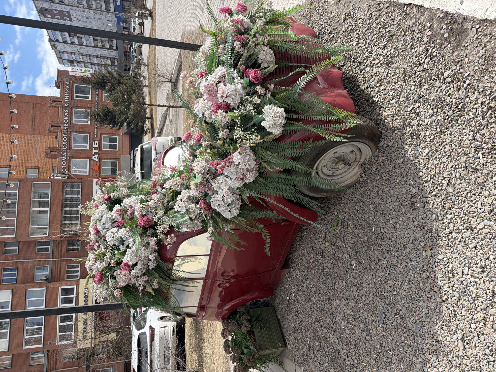 The image captures an outdoor urban scene featuring a vintage red car repurposed as a decorative floral display. The car, parked on a gravel surface, is extensively covered with a vibrant arrangement of artificial flowers in shades of pink, white, and red, intertwined with green fern leaves. One of its wheels is visible on the right, showing an aged tire and hubcap. Below the car, near its side, a green planter box with foliage is also visible.
In the background, several buildings line what appears to be a street or commercial area. Prominently featured is a brown brick building with multiple windows. Visible text on this building includes "СТОМАТОЛОГИЧЕСКАЯ КЛИНИКА" (Dental Clinic) and "АТБ," likely referring to a local business or supermarket chain. Below "АТБ," faint Cyrillic text can be deciphered as "Азиатско-Тихоокеанский Банк" (Asia-Pacific Bank). Taller, multi-story residential or office buildings are visible further in the distance. Streetlights with exposed bulbs are on the left, suggesting a commercial or pedestrian zone.
The scene is illuminated by bright daylight, indicating a clear, sunny day. Shadows are visible on the gravel and parts of the car. No people are explicitly visible in the frame, but the urban setting and decorative car imply human presence and activity. The overall impression is one of an artful urban embellishment. The location context provided confirms this is in Krasnoyarsk, Russia.