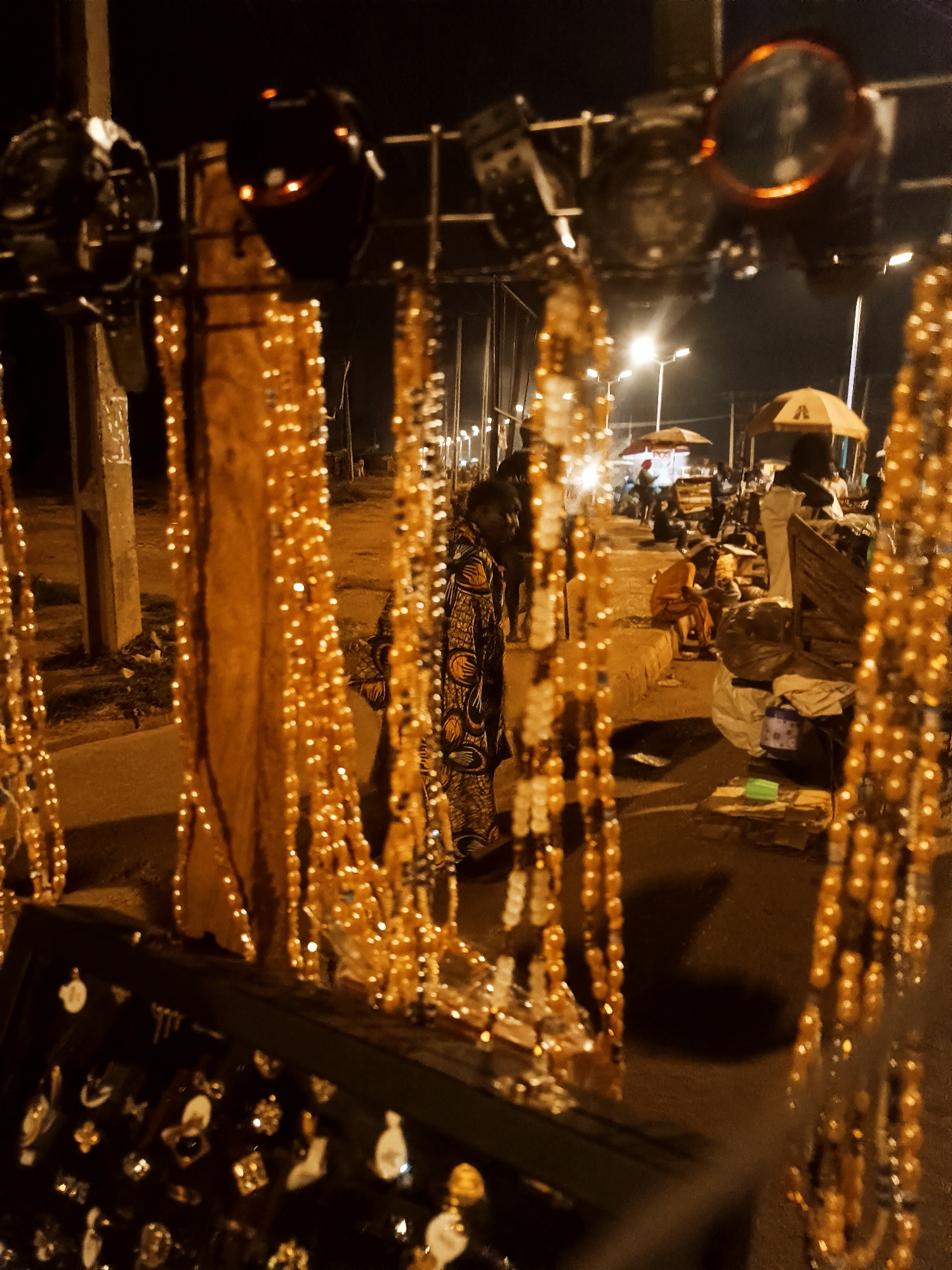 The image captures a nighttime scene in Bukuru, Nigeria. In the foreground, an assortment of jewelry, including necklaces and earrings, are displayed on a stand. The jewelry stand appears to be part of a street vendor's setup. Behind the jewelry, several people are visible in the background. Some stand near stalls or tables, while others sit or are engaged in conversation. Streetlights illuminate the area, casting a warm glow over the scene. The setting suggests a bustling night market or street fair in Bukuru. The focus is drawn to the jewelry, suggesting it is the primary subject of the photo.