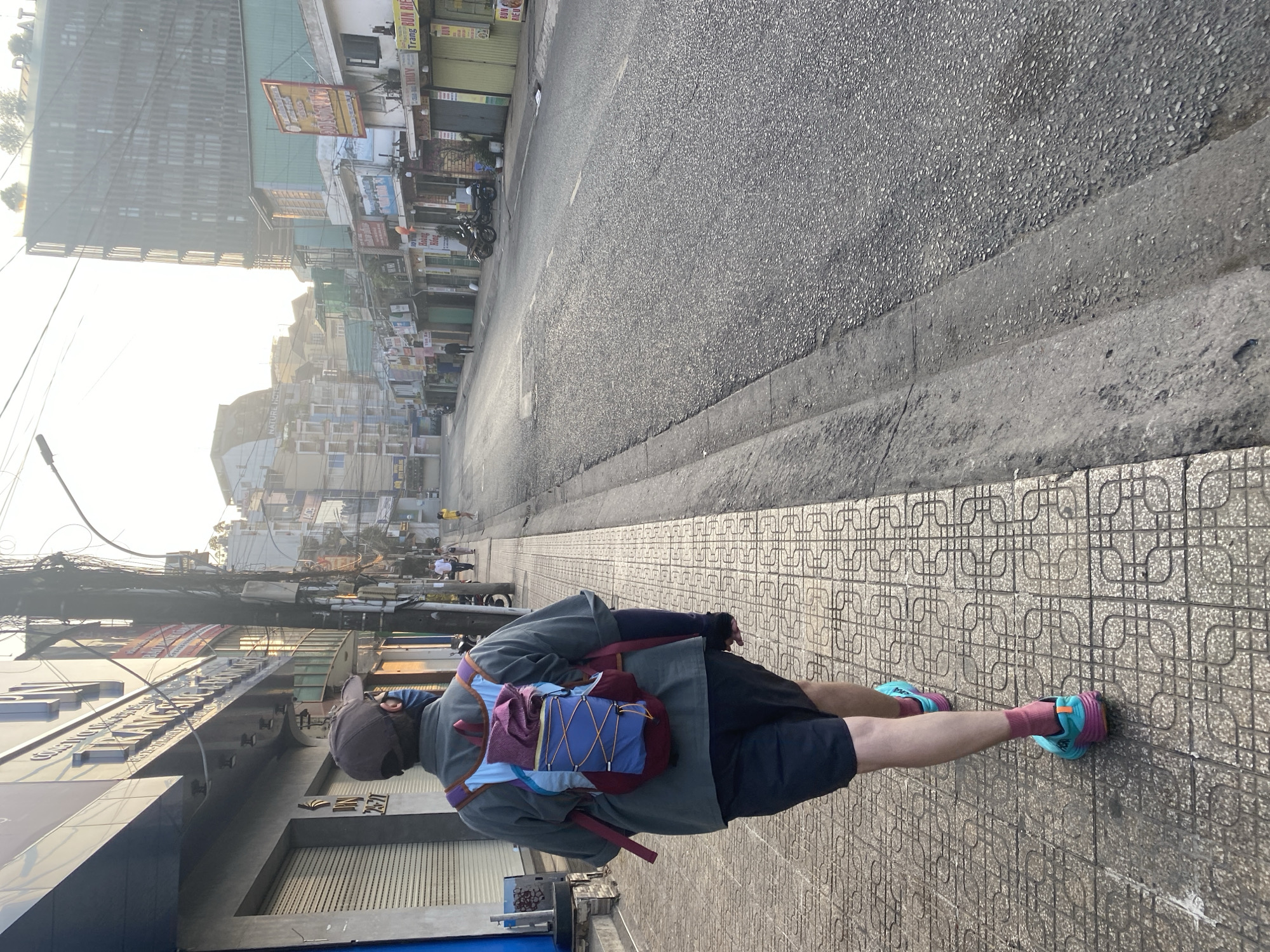 A person wearing a gray shirt, black shorts, a dark-colored cap, pink socks, and teal and pink running shoes is walking away from the viewer on a tiled sidewalk. The person is wearing a backpack with maroon, blue, and lavender panels. The sidewalk is adjacent to a road, which is bordered by buildings. In the distance are more buildings, and overhead is a network of electrical wires. The photograph was taken in Da Lat, Vietnam.