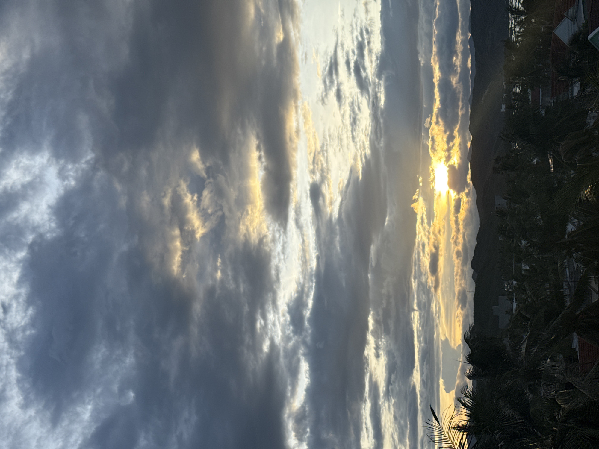 A wide-angle, high-angle shot captures a sunset over a tropical landscape. The sky is filled with dramatic, dark gray clouds, with patches of bright yellow sunlight breaking through. The sun itself is visible as a bright, hazy orb, partially obscured by clouds, casting rays of light downwards. Below the sky is a dark silhouette of rolling hills and mountains. In the foreground, the fronds of palm trees are silhouetted against the sky, their leaves appearing windblown. Below the palm trees, a scattering of rooftops and buildings can be seen, suggesting a coastal or resort town. The overall impression is one of a dramatic and slightly moody natural scene at dusk.