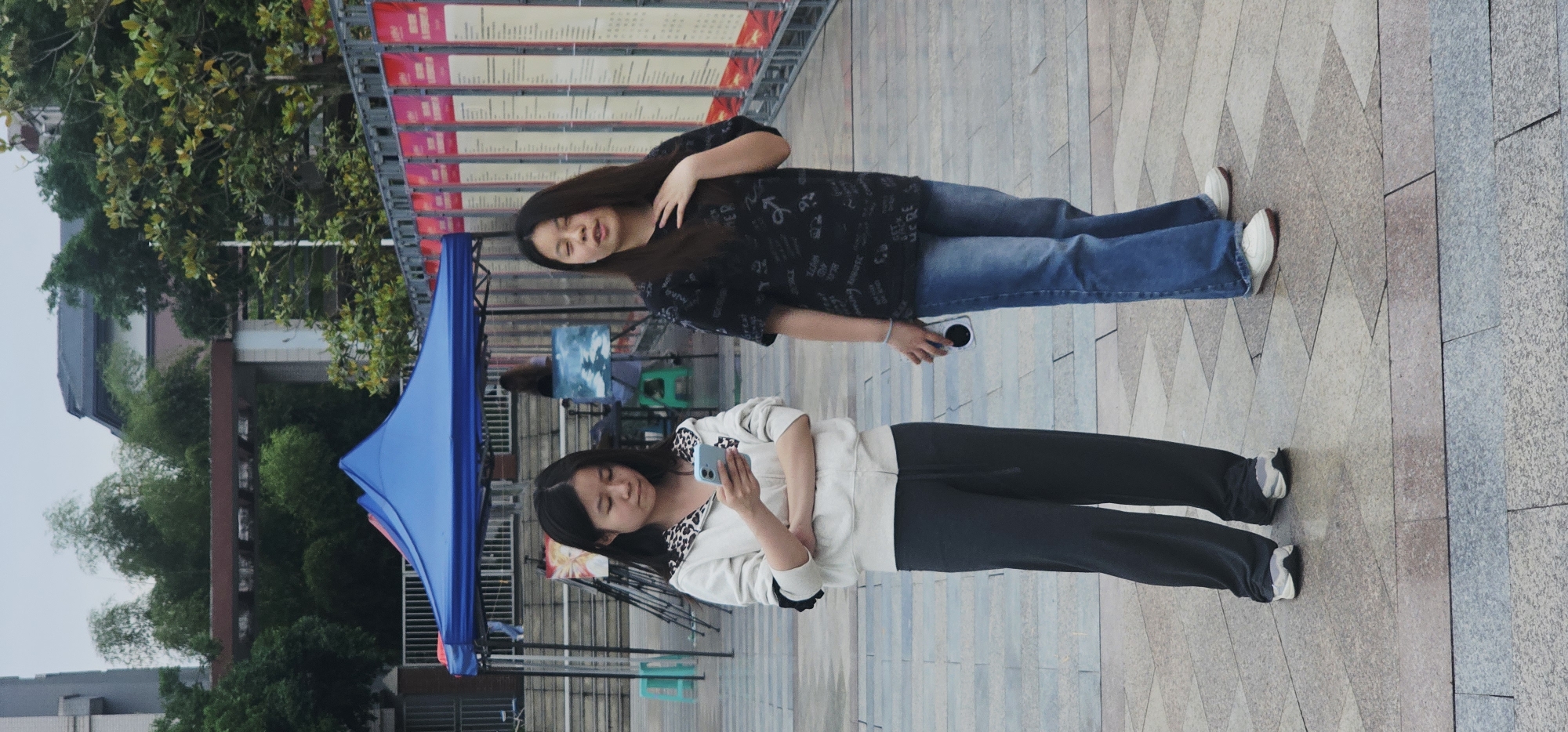 Two young women stand on a patterned stone walkway. One woman, wearing jeans and a patterned black t-shirt, has long dark hair and is smiling while holding a small white object. The other woman, wearing dark sweatpants and a white hoodie, is looking at her cell phone. A blue tent stands behind the woman with the cell phone. In the background are trees and a building. A red and white banner is visible above the woman in jeans. The image appears to be taken in a public area in Chongqing High-tech Zone, China.