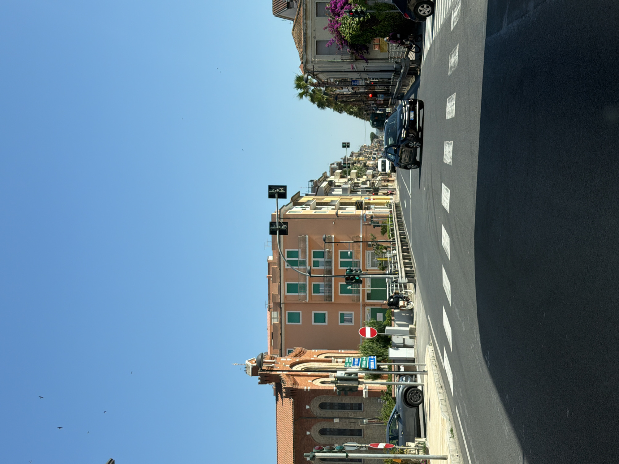 The image depicts a street scene in Potenza Picena, Italy, on a clear day. A wide, paved road occupies the foreground, with a pedestrian crossing marked by white stripes. To the left of the road, a grey car is parked on a sidewalk next to a building with a red-tiled roof and arched windows, suggesting a church or similar institutional structure. Traffic signals are visible at an intersection.
Further down the street, buildings of various heights and colors line both sides. The buildings feature balconies and windows, with some adorned with greenery. Several cars are on the road, including a black SUV in the middle lane and a white van in the distance. A person is riding a bicycle on the right side of the road. Palm trees are present along the right side of the street, adding a tropical element to the urban landscape. The sky is a clear blue with a few birds in flight. The overall impression is of a typical town street with moderate traffic and pedestrian activity.