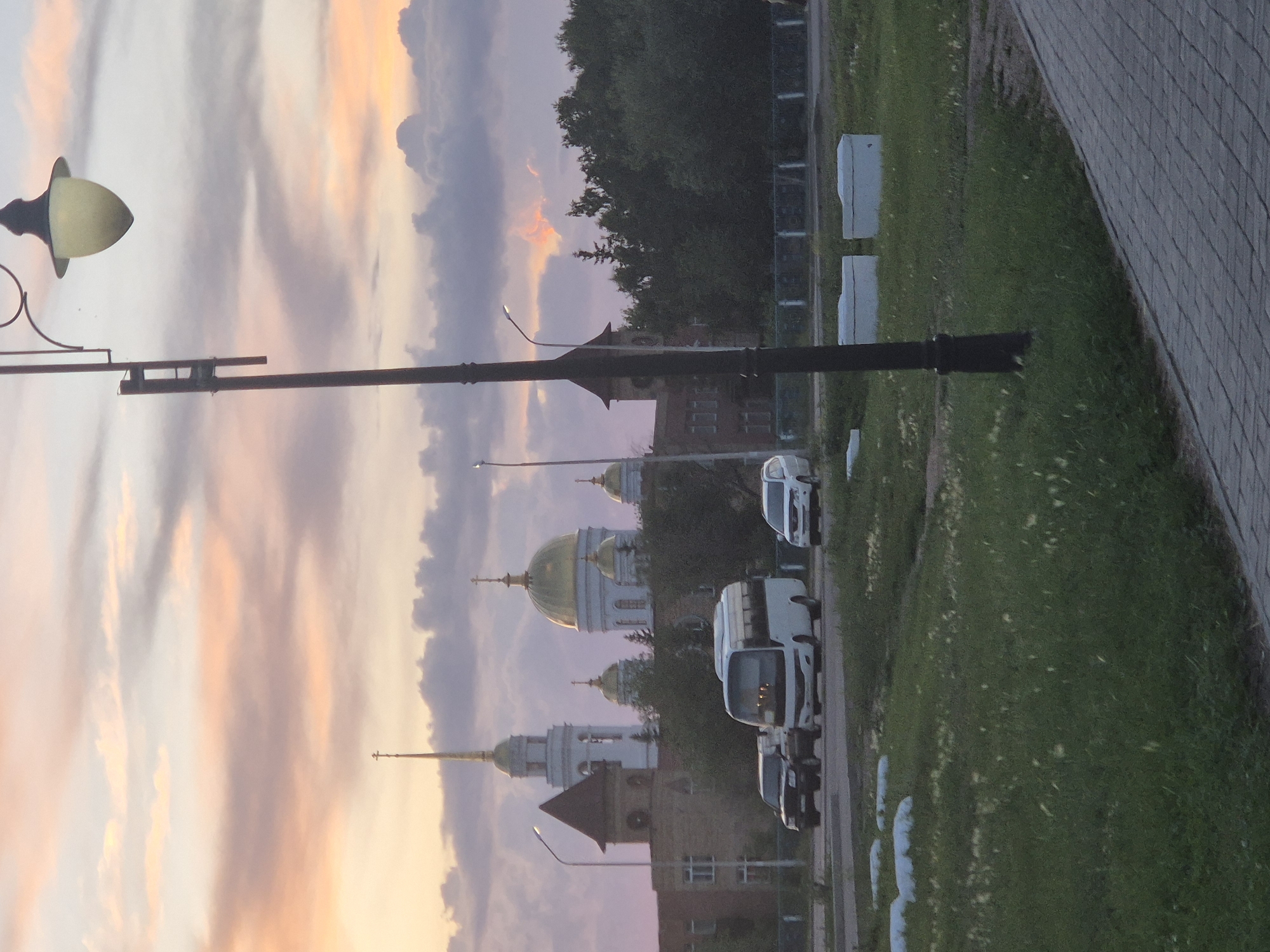 The image shows a street scene in Kokshetau, Kazakhstan, at sunset. In the background, a cluster of buildings with golden domes and spires suggests a religious complex, possibly a cathedral or monastery. A prominent lamp post stands in the foreground, casting a shadow. Several vehicles, including a white bus and two cars, are visible on the street. The sky is filled with dramatic clouds in shades of pink, orange, and purple, indicative of the setting sun. A paved walkway runs along the right side of the image, and a grassy area with some white objects, possibly landscaping elements, is present in the foreground. The overall atmosphere is serene and picturesque, capturing a moment of twilight in an urban setting.