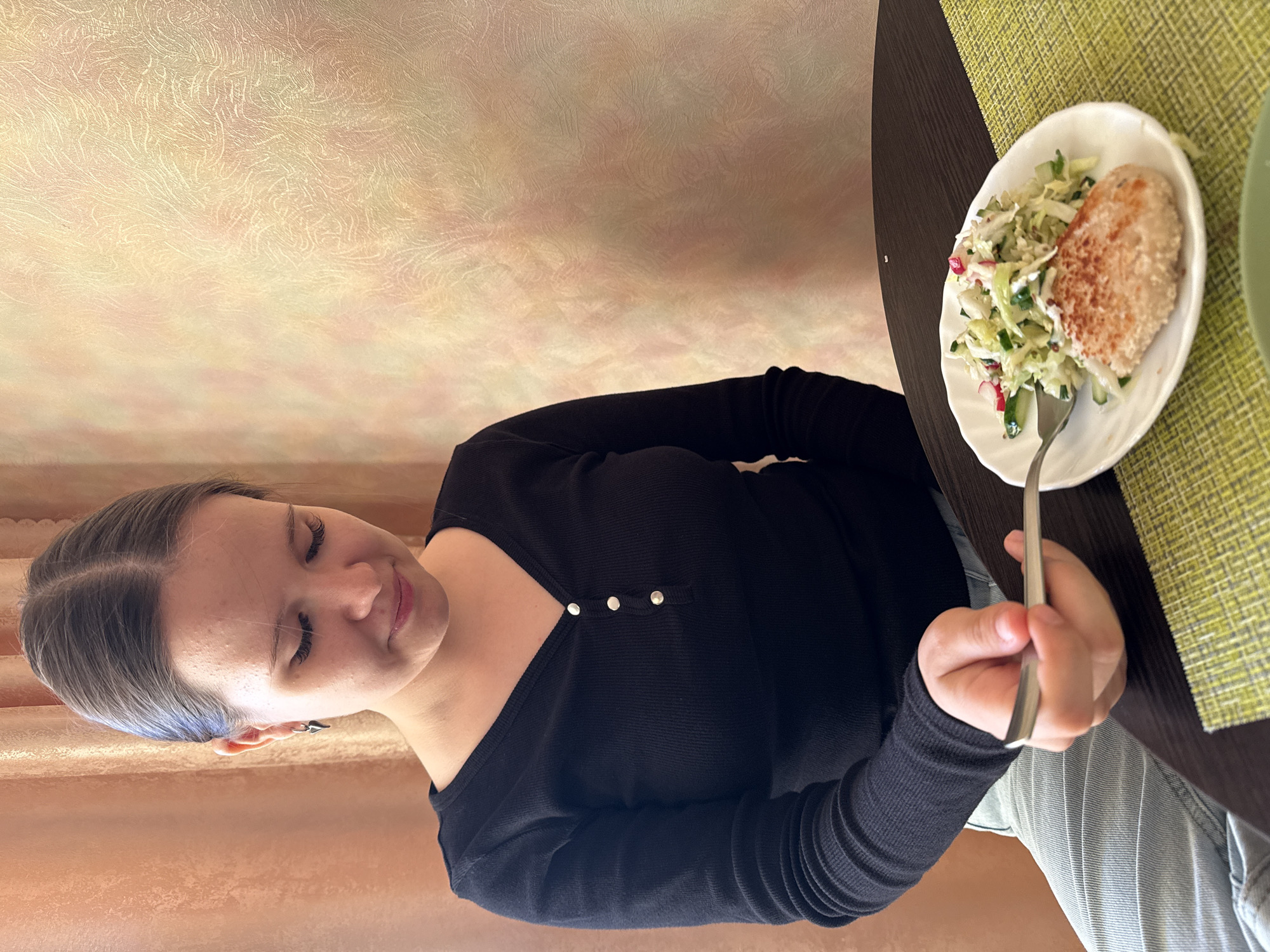 A young woman with light-colored hair, styled with a middle part and pulled back, is seated at a dark brown table, enjoying a meal. She wears a black long-sleeved top featuring three small silver buttons near the neckline and light blue trousers or jeans. A subtle, dark, spike-shaped stud earring adorns her left ear. Her gaze is directed downwards towards her plate, and a soft smile suggests contentment or focus on her food.
In front of her, on a light green textured placemat, rests a white, scalloped-edge plate. The plate holds a meal comprising a fresh salad made with chopped green vegetables (likely cabbage or lettuce and cucumber) and pieces of red vegetable (possibly radish), alongside a golden-brown breaded patty, possibly a cutlet or fish cake. A silver fork is held in her right hand, poised within the salad.
The setting is indoors, with a textured wall featuring light yellow and orange tones visible in the background, resembling wallpaper. The atmosphere appears casual and calm. There is no discernible text in the image. The scene captures a moment of dining in an indoor setting, likely a home in Izhevsk, Russia.