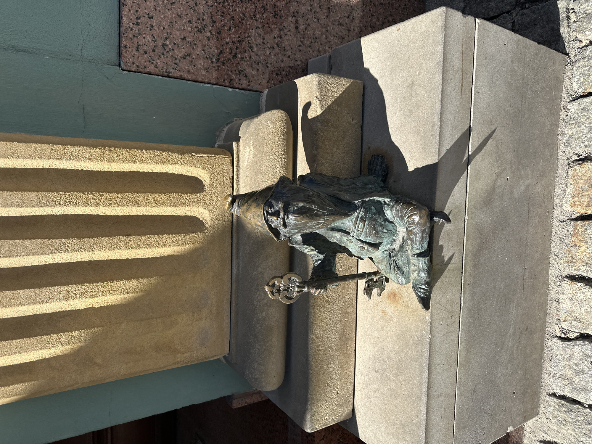 A bronze dwarf statue clings to the side of a stone building facade in Wrocław, Poland. The dwarf is holding a key. The stone facade features horizontal grooves and varying stone textures. The scene is illuminated by strong sunlight.