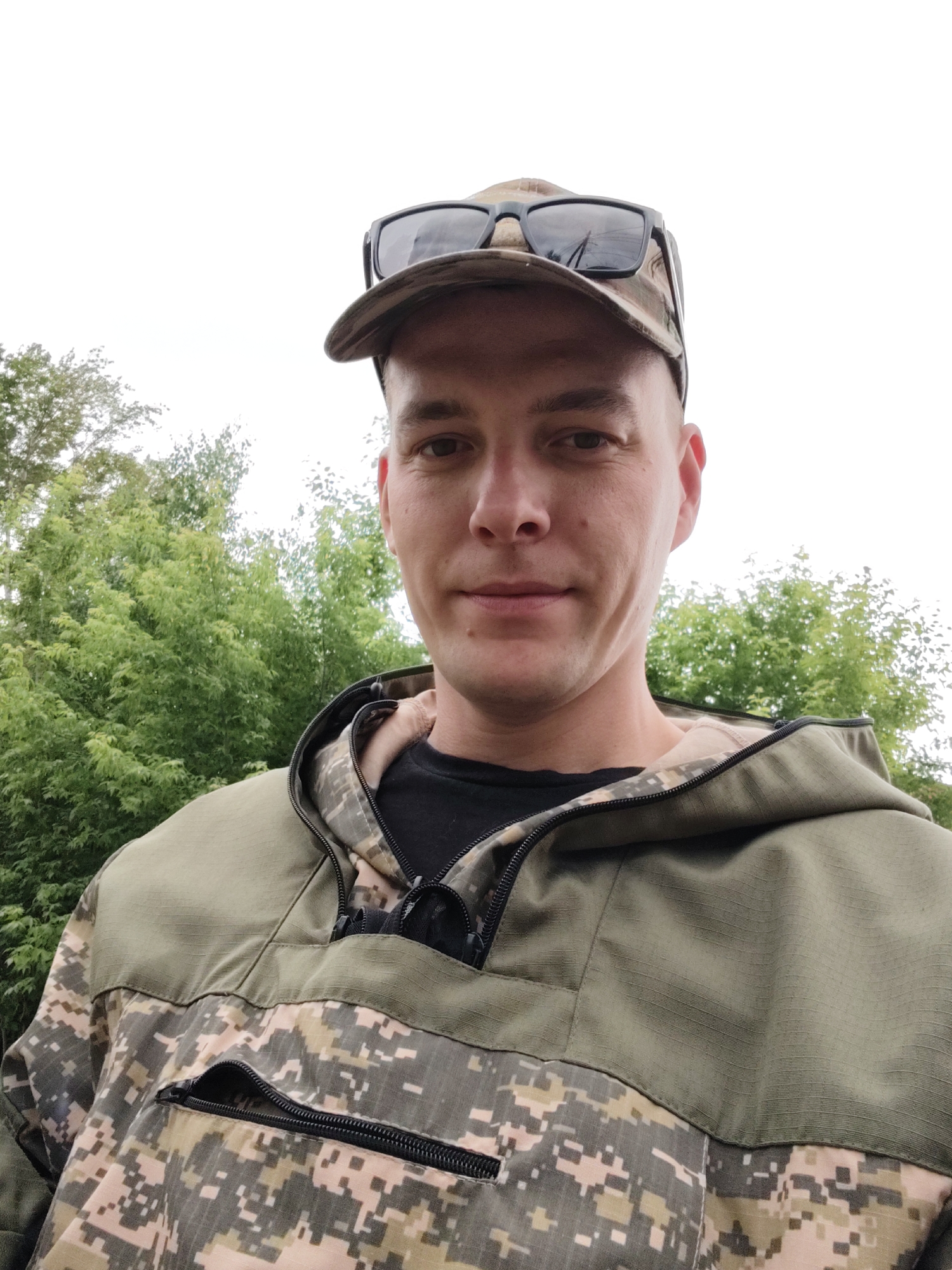 A medium shot shows a man from the chest up, wearing a camouflage jacket and a camouflage baseball cap with sunglasses resting on top. He is outdoors, with lush green trees forming the background under a bright, overcast sky. The man has short brown hair, fair skin, and appears to be looking directly at the camera with a neutral expression. The jacket has a zipper at the neck and a zippered pocket on the front. The overall impression is that of a casual, outdoor portrait taken in a natural environment. The image is not specific to Chaikovsky, Russia, as no identifying landmarks or contextual clues are present.