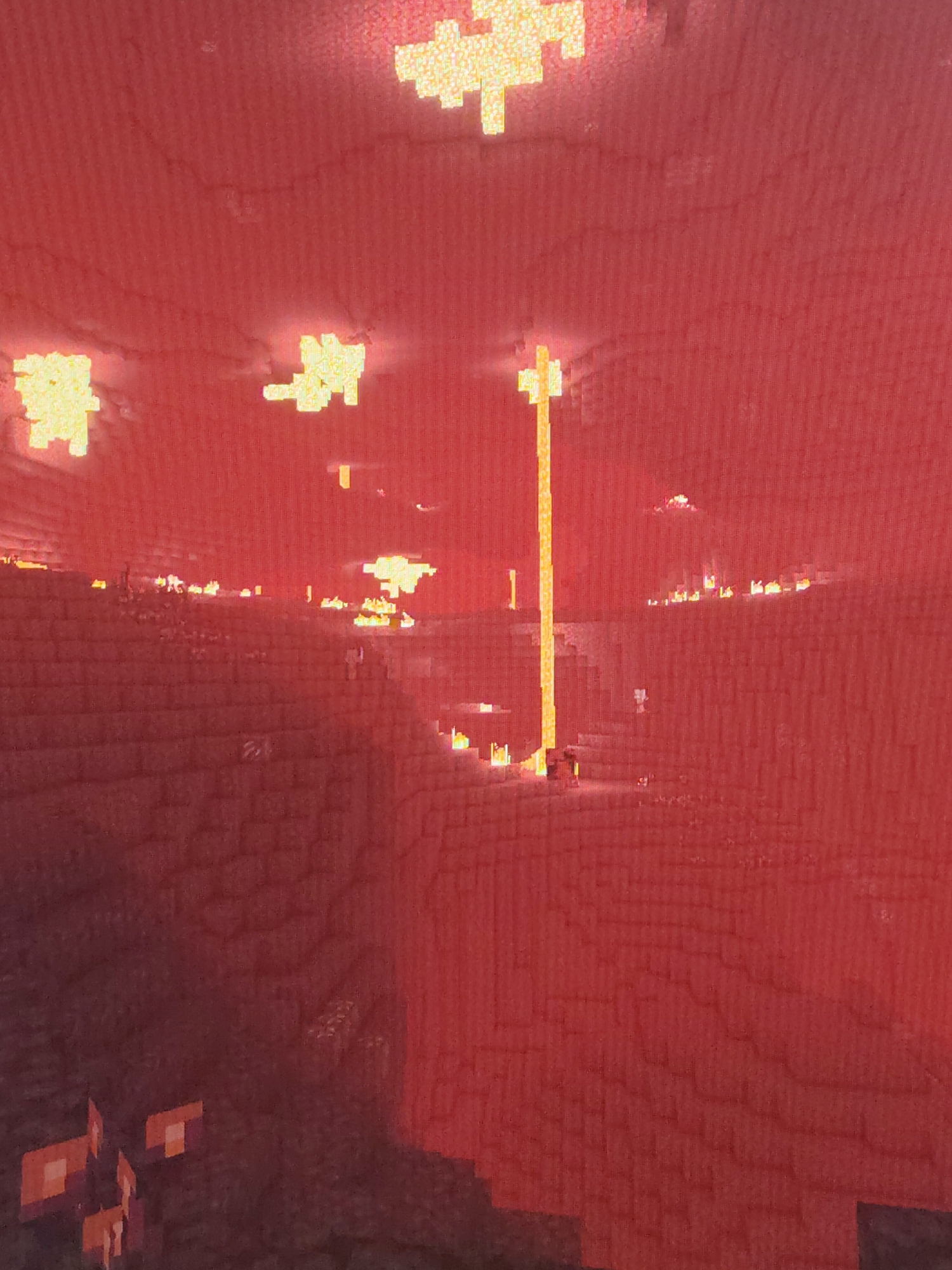 The image depicts a scene from the video game Minecraft, specifically the Nether dimension. The environment is predominantly red, with blocks of netherrack forming the ground and walls. The ceiling is also netherrack and features scattered patches of light sources, possibly glowstone. A prominent pillar of lava rises from the ground. In the foreground, there are nether wart blocks. The overall impression is of a dark, fiery, and cavernous landscape. There are no people or subjects present.