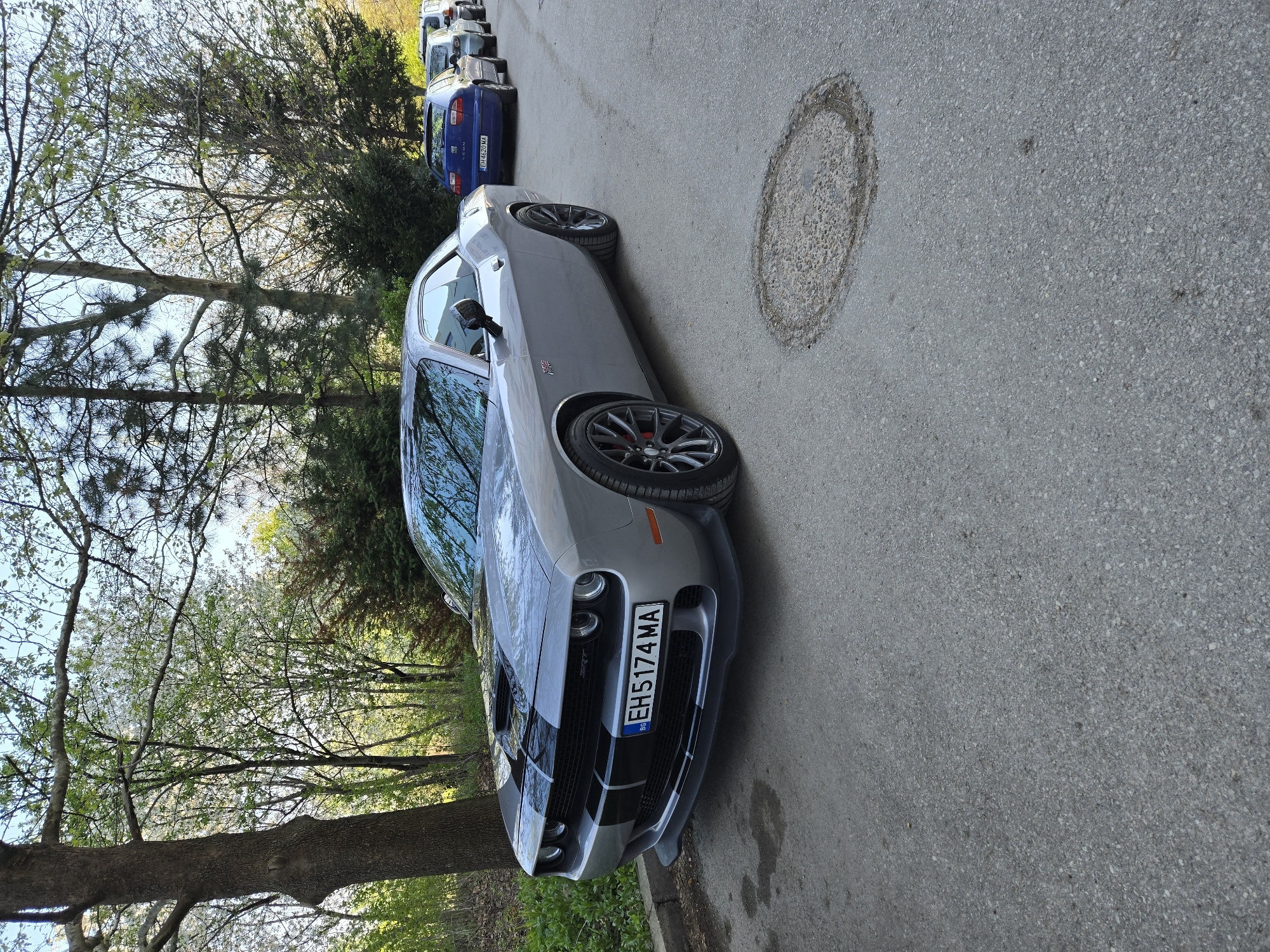 A gray Dodge Challenger with the license plate "EH5174MA" is parked on a paved street in an urban or suburban environment. The car is positioned on the right side of the road. In the background, other vehicles are parked. The scene includes trees and vegetation. There is a manhole cover visible on the road. The specific location within the city of Unknown, Unknown is not discernible from the image.