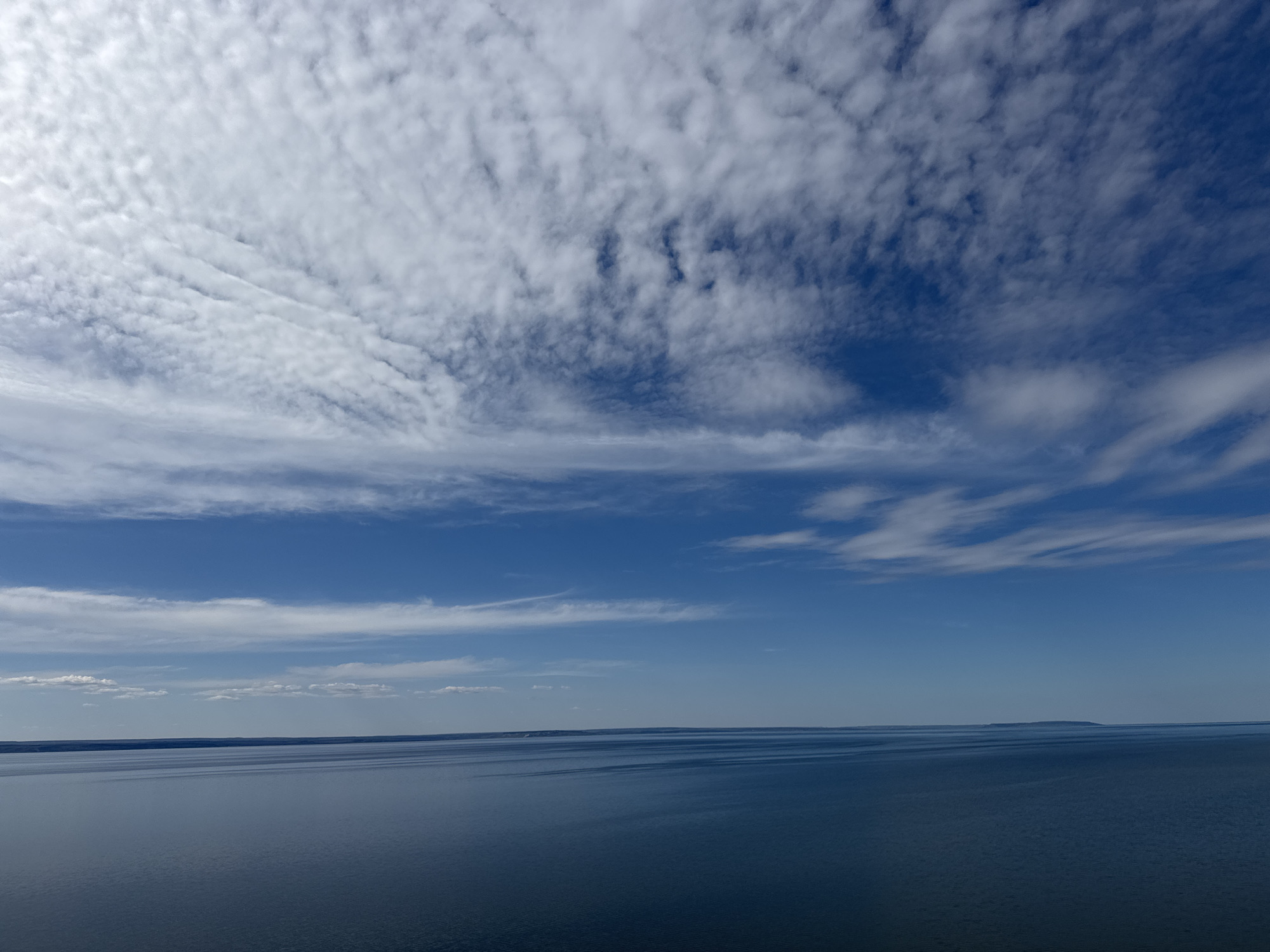 The image depicts a body of water, possibly a lake or ocean, under a blue sky with scattered clouds. The water appears calm and reflects the sky. In the distance, a faint landmass or island is visible on the horizon. The location is unknown. There are no people, subjects, objects, actions, or interactions readily apparent in the image beyond the natural elements of water, sky, and clouds.