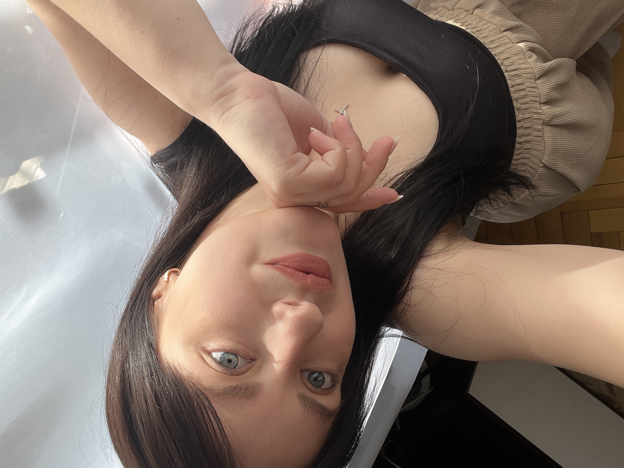 A selfie taken by a woman with dark hair and light eyes. She is wearing a black top and beige pants. Her hand is near her face, and she is looking at the camera. The location is likely indoors, but the specific details cannot be determined from the image, nor can a specific city or address be determined.