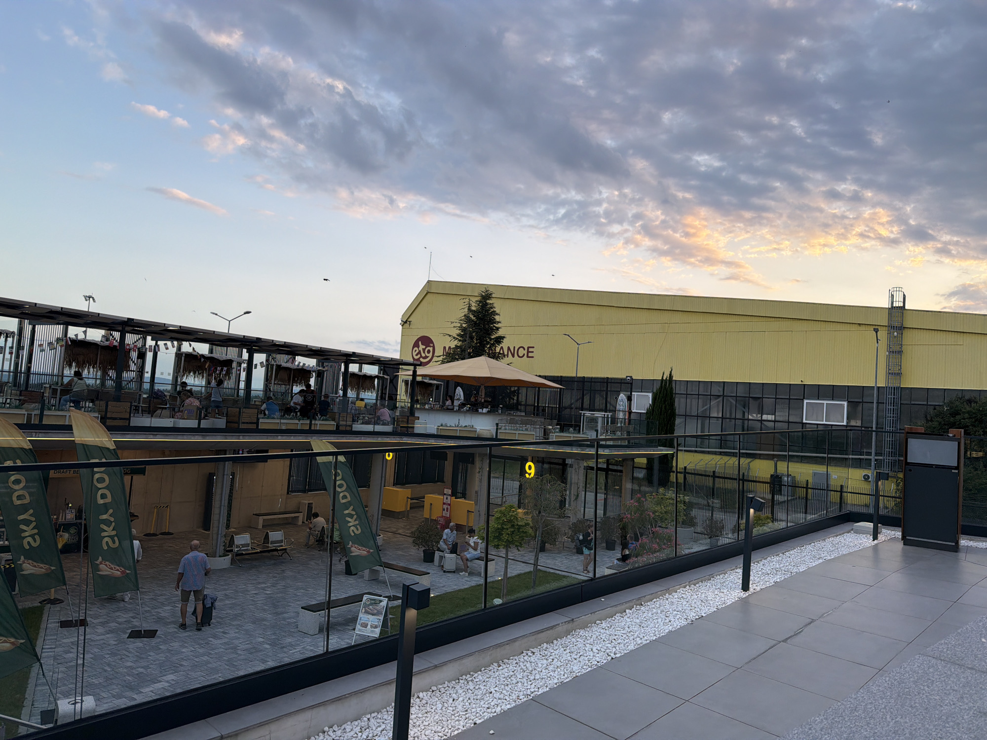 The image captures an outdoor area with a mix of modern and rustic elements under a cloudy sky tinged with sunset colors. In the background, a large, bright yellow industrial-style building dominates the skyline. A sign on the building reads "ETG MAINTENANCE". In the mid-ground, a covered structure with thatched roofing and exposed wooden beams provides seating for patrons, suggesting a restaurant or bar area. An orange umbrella shades a portion of this area. Below this, a paved courtyard is visible, bordered by a glass partition. Several green flags with white text and images of hot dogs are planted in the ground. A man in a patterned shirt and shorts is standing in the courtyard, facing away from the camera, with a small wheeled suitcase at his feet. Other individuals are seated at tables and on benches within the courtyard and in the background seating area. The foreground features a paved walkway with a decorative border of white pebbles and several modern light fixtures. The overall scene suggests a public gathering space, possibly a commercial area or a venue, located in Aksakovo, Bulgaria.