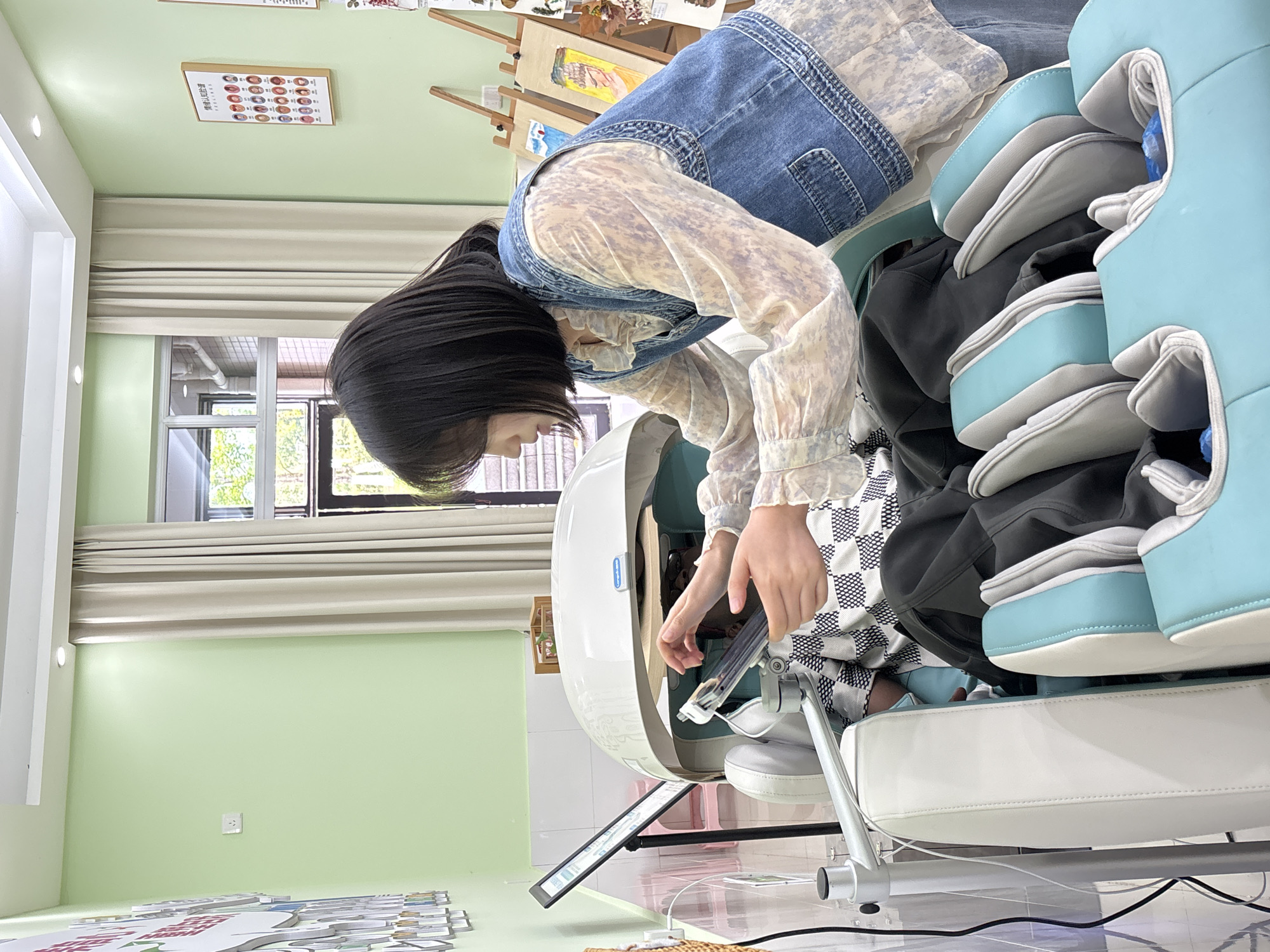 A medium shot captures a young Asian woman as she interacts with a complex piece of equipment, likely a diagnostic or therapeutic device, situated in an indoor setting in Jiangjin, China. The woman has black hair and is wearing a long-sleeved, light-colored shirt with a denim vest. She is bending over and appearing to manipulate part of the equipment with her hands. The equipment is largely white with light blue accents. It appears to have a screen or display panel, possibly indicating digital functions. The surrounding environment includes a window with natural light filtering in, curtains, and pale green walls. A poster with an array of faces is affixed to the wall. An easel and painting supplies are visible in the background. The scene appears to be indoors, possibly a clinic, rehabilitation center, or school-related facility.