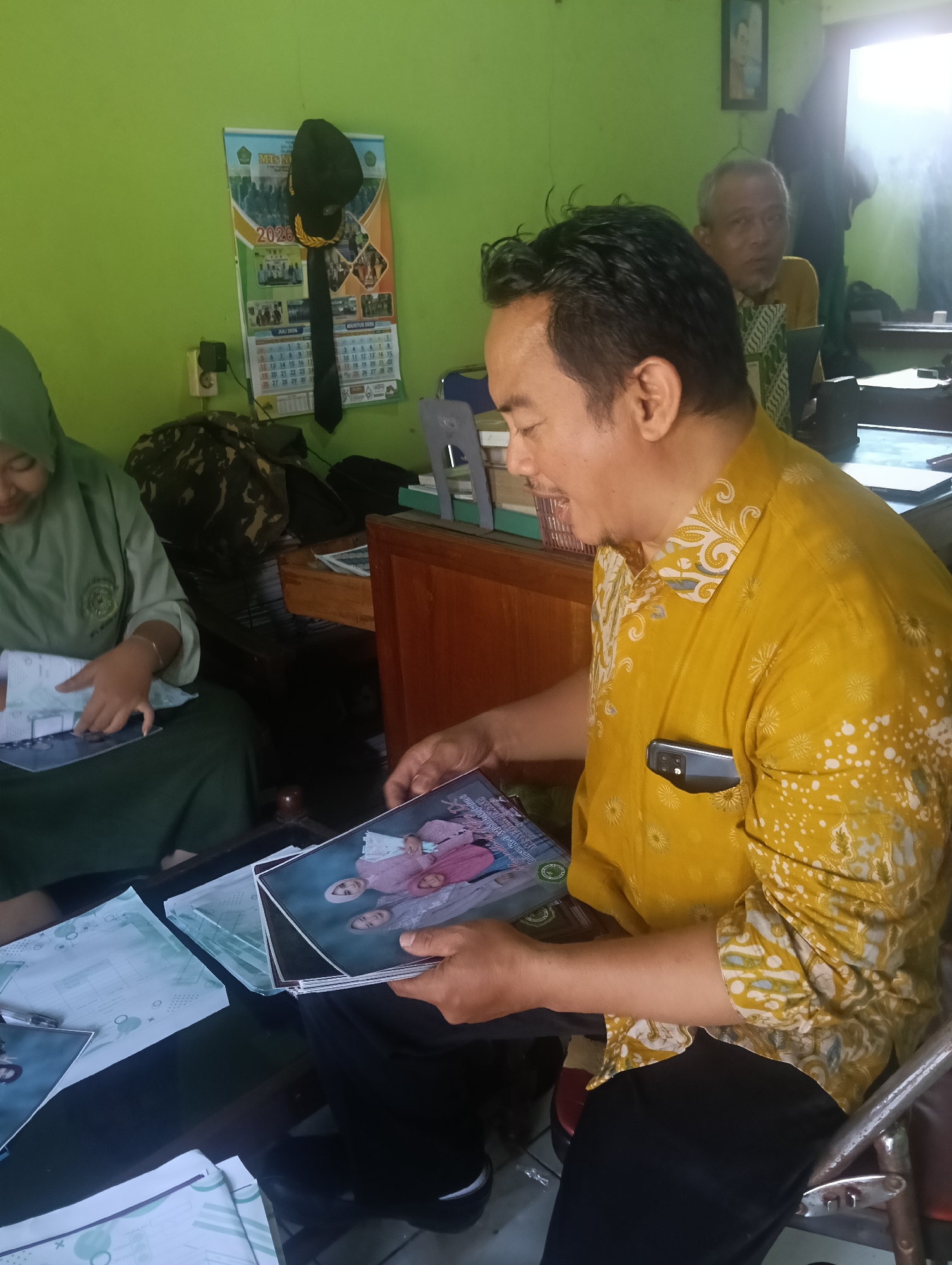 In the city of Adinuso, Indonesia, a man wearing a yellow batik shirt is seated and looking down at a stack of photographs. A woman in a green hijab and uniform is seated to his left, also looking at documents. In the background, an older man with glasses is visible. A calendar for 2024 hangs on a green wall. The scene appears to be an indoor office setting.