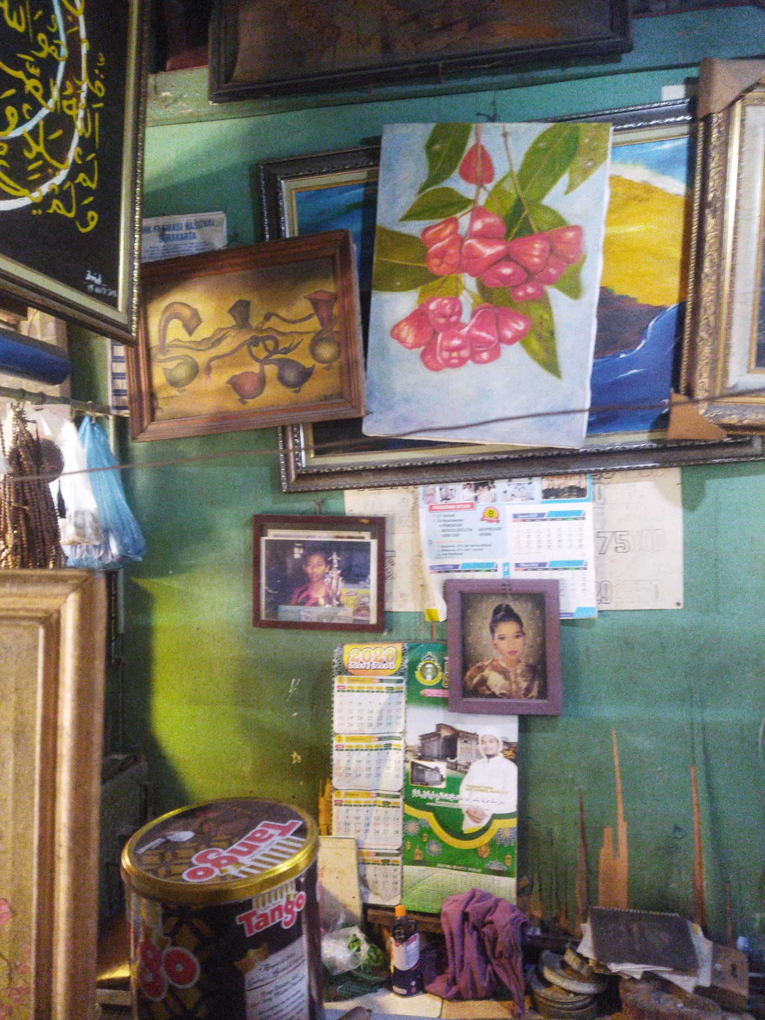 The image depicts a densely cluttered wall in a room, possibly a shop, in Surakarta, Indonesia. The wall is painted a faded green and serves as a backdrop for an array of framed pictures, calendars, and miscellaneous objects.
Dominating the upper left is a large framed artwork with yellow Arabic calligraphy on a black background. To its right, a painting of red, bell-shaped fruits hangs prominently. Below these, a smaller framed painting shows a still life with stylized fruit shapes.
Several framed photographs are displayed at various heights. One shows a young woman in a red top. Another, a more recent portrait, features a young woman with dark hair pulled back, wearing traditional attire. A framed image of a mosque and a man in white attire is also visible.
Calendars are interspersed among the framed items, with at least one identified as "2020". These are stacked and overlapped, adding to the layered appearance of the display.
Hanging to the left of the framed artworks are brown beaded necklaces or rosaries, along with two plastic bags, one white and one blue.
In the foreground, a cylindrical tin labeled "Tango" is visible. It appears to be a food or drink product. Scattered around this are various other objects, including what seem to be tools, piles of paper, and a folded piece of dark purple fabric. The overall impression is one of a lived-in, possibly commercial space, filled with personal and decorative items. The lighting suggests an indoor environment, possibly with natural light contributing to the shadows.