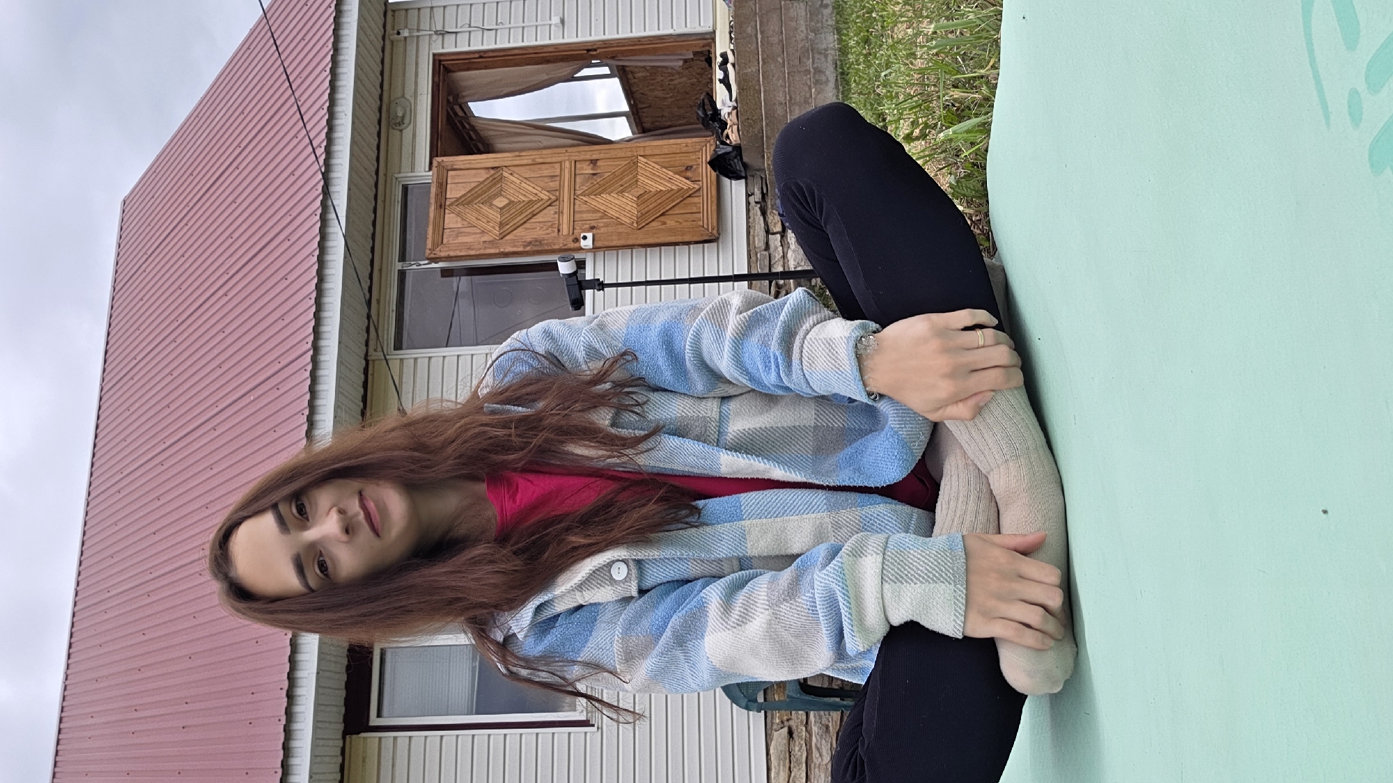 A young woman with long brown hair is sitting cross-legged on a light green mat outdoors. She is wearing a bright pink t-shirt and a blue and white plaid jacket. Her legs are crossed in front of her, and she is wearing light brown socks and black pants. Her hands are resting on her knees. Behind her is a light-colored house with a red metal roof. The house has a wooden double door that is open, revealing a dark interior. A black tripod with a camera on top is positioned to the left of the doorway. The ground is covered in green grass, and there are some stones and a plastic chair visible to the left of the woman. The sky is overcast.