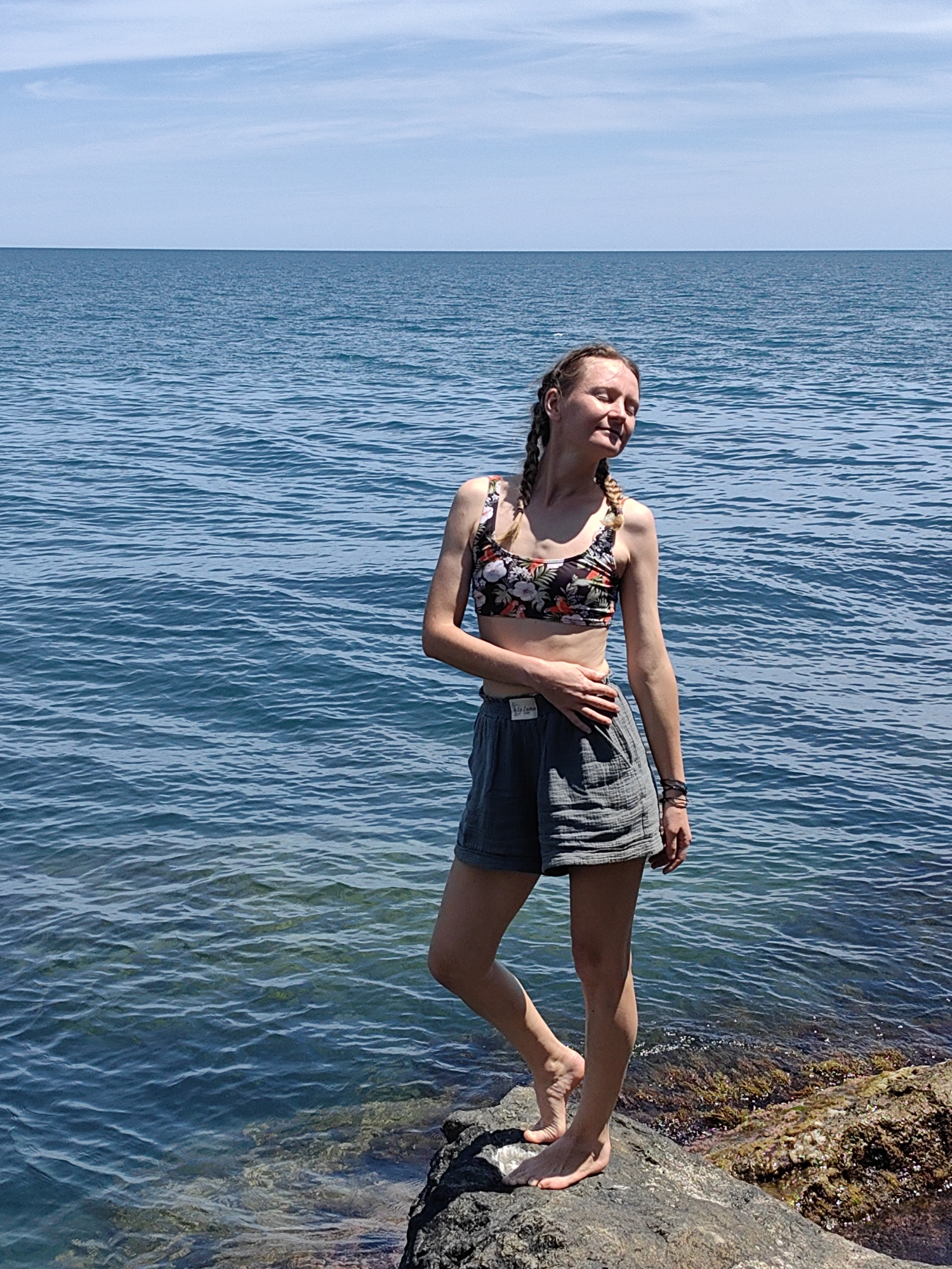 A young woman stands on a rocky shore, with her back to the sea, facing towards the right of the frame. She has fair skin and blonde hair styled in two braids. She is wearing a floral print bikini top and grey shorts. Her right hand rests on her hip, and her left hand is on her stomach. Her eyes are closed, and her head is tilted back slightly, suggesting she is enjoying the sun. The sea is calm and blue, stretching out to the horizon under a clear sky. The rocks in the foreground are dark and textured, with some patches of seaweed visible in the water. The location is the coast of Utios, Ukraine.