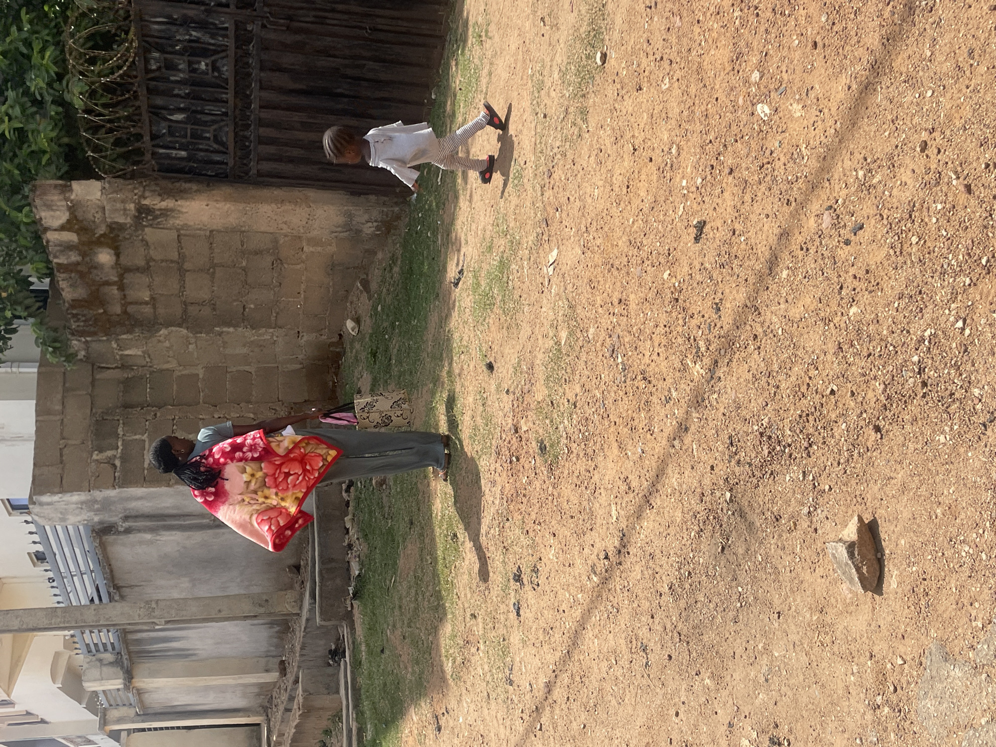 In Jos, Nigeria, an outdoor scene shows a woman and a child. The woman stands near a building, carrying a floral blanket and a patterned handbag. The child, dressed in light-colored clothing, is further out on a dirt and grassy area. A partial view of a fence with barbed wire is visible near the child. The ground is mostly dirt, with patches of grass. A stone or object is on the ground in the foreground. Buildings surround the scene.