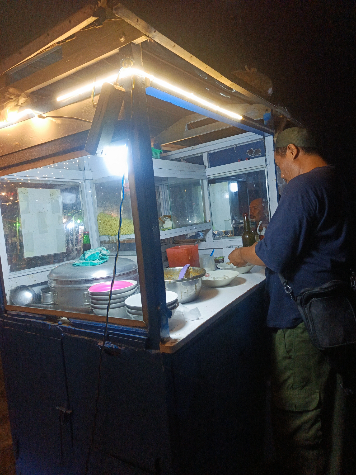 The image depicts a man preparing food at a food stall in Luwoo, Indonesia. The stall is illuminated by overhead fluorescent lights, casting a warm glow on the scene. The man, wearing a green cap and a dark blue t-shirt, is seen working with bowls and utensils, suggesting he is in the process of serving a customer.
The stall itself is made of wood and glass, with various food items and cooking equipment visible. A large metal pot sits on a shelf, likely containing a stew or soup. A stack of plates and bowls are neatly arranged, ready for use. In the background, there are glimpses of other food items, including what appears to be noodles in a container. The overall impression is one of a bustling street food vendor, providing a meal to the local community. The time of day appears to be evening or night, given the artificial lighting and the dark background.
