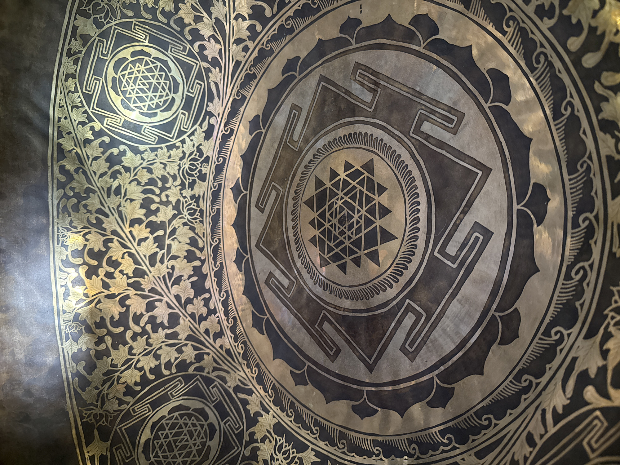 A close-up shot focuses on a metallic gong-like object adorned with geometric and floral designs. The central design consists of concentric circles enclosing a pattern of intersecting triangles. Around this design are stylized floral and geometric elements that appear to be etched or inlaid into the metal. The lighting highlights the metallic sheen and textures of the surface. No people or specific environmental details are visible, so the location in Tashkent, Uzbekistan, cannot be independently verified from this image alone.