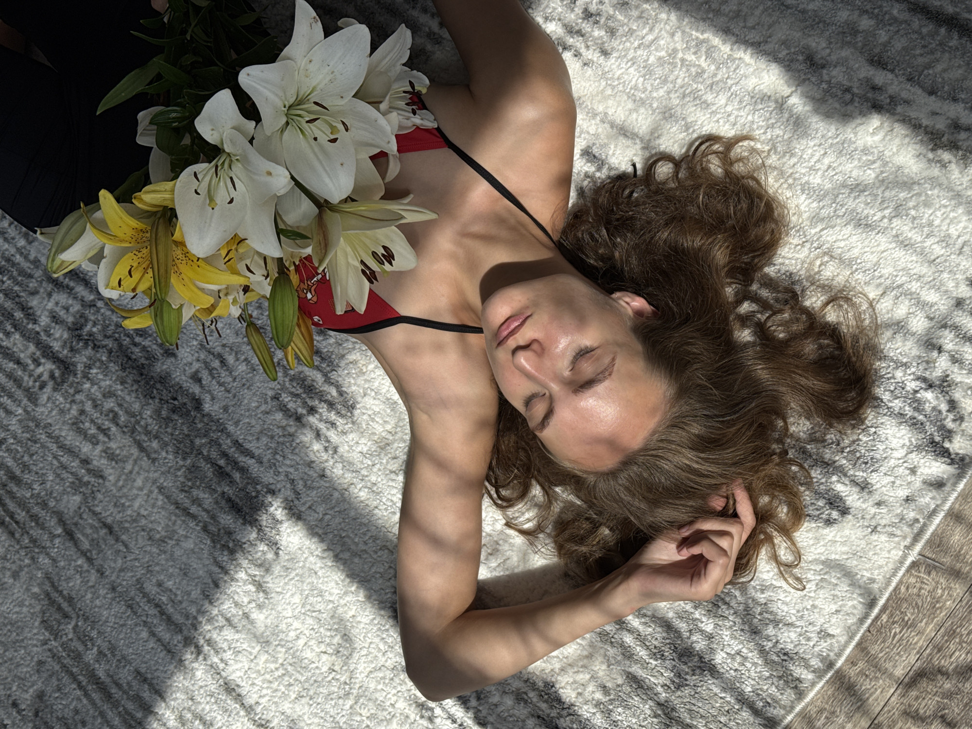 In Sharkan, Russia, a person lies on a rug with a bouquet of lilies on their chest. The person is wearing a red bikini top with black straps. Their eyes are closed, and their head is tilted back. Sunlight casts shadows on the rug and the person's body. The environment appears to be indoors, with a wooden floor visible to the left of the rug.