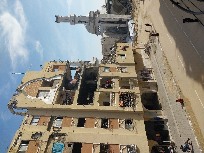 The image depicts an urban street scene in Khan Yunis, Palestinian Territory, characterized by extensive structural damage under a partly cloudy sky.
In the left half of the frame, a multi-story, beige-colored building, likely residential or commercial, exhibits severe damage. Large sections of its facade and internal floors are missing or collapsed, exposing interiors, rebar, and debris. The exterior walls are pockmarked with numerous small impact holes. Architectural features such as arched window surrounds are visible on the less damaged sections. Clothes hang from at least two balconies, and a blue tarp is visible from an upper-floor window. At ground level, an open storefront displays clothing on racks.
In the background to the right, a white mosque with a tall, slender minaret and a dome is partially visible. The mosque also shows signs of damage, particularly around the dome and minaret's upper sections, with rubble visible at its base.
A two-lane asphalt road with a double white centerline runs horizontally across the mid-ground. A paved sidewalk borders the road, transitioning into sandy, debris-strewn areas. Piles of rubble are scattered across the scene, particularly between the damaged building and the mosque, where white tarpaulins cover areas of debris or possibly makeshift structures.
Eight individuals are present. On the sidewalk near the damaged building, two people are seated on chairs next to the clothing display. A single person in red clothing sits on the curb further down the street. Three individuals walk through the sandy, debris-laden area towards the mosque, with one appearing to carry an item. One person in dark clothing walks on the sidewalk. Another person, also in dark attire, is partially visible in the bottom right foreground, walking on the road.
