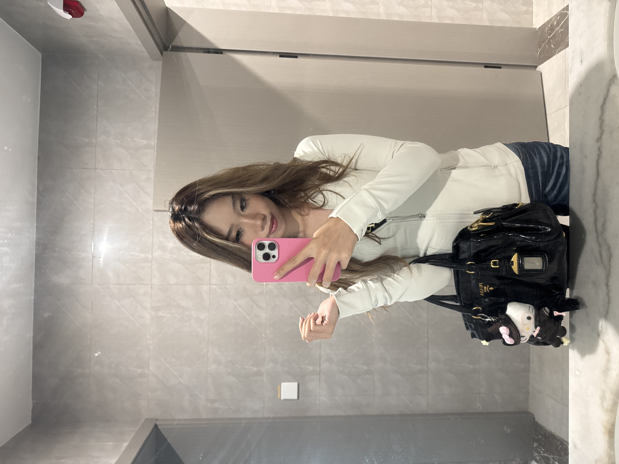 A woman is taking a selfie in a bathroom with gray marble walls, likely in Nan'an District, China. She is holding a pink smartphone with her right hand. She's wearing a white zippered jacket and blue jeans. A black patent leather handbag with gold hardware and a Hello Kitty keychain is beside her. The bathroom contains a large mirror, a light switch, and potentially a shower or door.