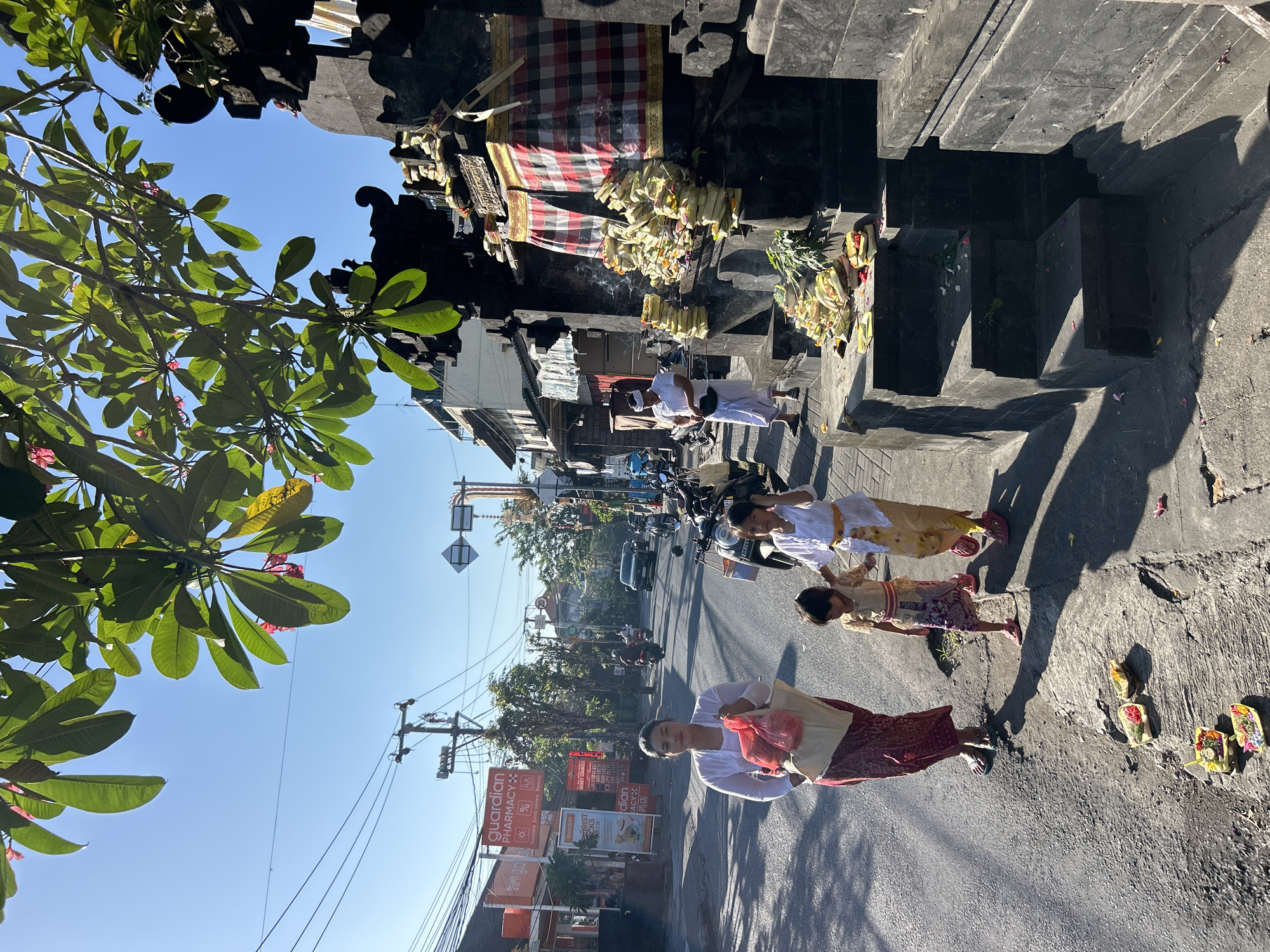 This image depicts a street scene in North Kuta, Indonesia, bathed in the bright sunlight of what appears to be late morning or early afternoon. The perspective is from street level, with a tree in the foreground on the left casting dappled shadows. In the background, the scene is dominated by a dark stone structure, possibly part of a temple or religious site, adorned with offerings. These offerings, primarily composed of woven palm leaves filled with flowers and possibly food, are also scattered on the side of the road.
Several people are visible. On the right side of the road, a woman in traditional Indonesian attire, a red and white patterned skirt and a white top, is walking away from the camera, carrying a light-colored bag. She appears to be smiling. Further up the street, three children, two girls and a boy, are walking in the same direction. The girls are dressed in colorful traditional clothing, including one in a yellow and white outfit and another in a yellow and patterned skirt. The boy is in a light-colored shirt. Their expressions are neutral. Another person in white attire is partially visible near the stone structure, possibly engaged in prayer or ritual.
The street itself is paved and shows signs of traffic, with a car and several motorbikes visible in the mid-ground. Buildings line the street, including what appears to be a pharmacy with signage that reads "GUARDIAN PHARMACY" and other commercial establishments. Power lines and utility poles are prominent, indicating an urban or semi-urban setting. The sky is a clear, bright blue, suggesting fair weather. The overall atmosphere seems peaceful and culturally rich, with the presence of religious offerings and traditional dress.