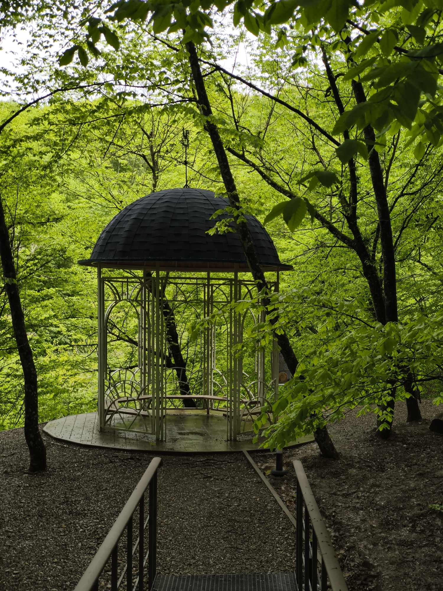A gazebo is located in a wooded area in the city of Goryachiy Klyuch, Russia. The gazebo is elevated on a circular platform covered in small pebbles and can be reached by a set of metal stairs with metal handrails. The gazebo itself has a dark-colored, dome-shaped roof and is surrounded by thin, light-colored metal posts. Inside the gazebo, there is a bench for sitting. The surrounding area is heavily wooded with vibrant green trees, some of which grow through the platform of the gazebo. The ground is covered with small pebbles and dirt.
