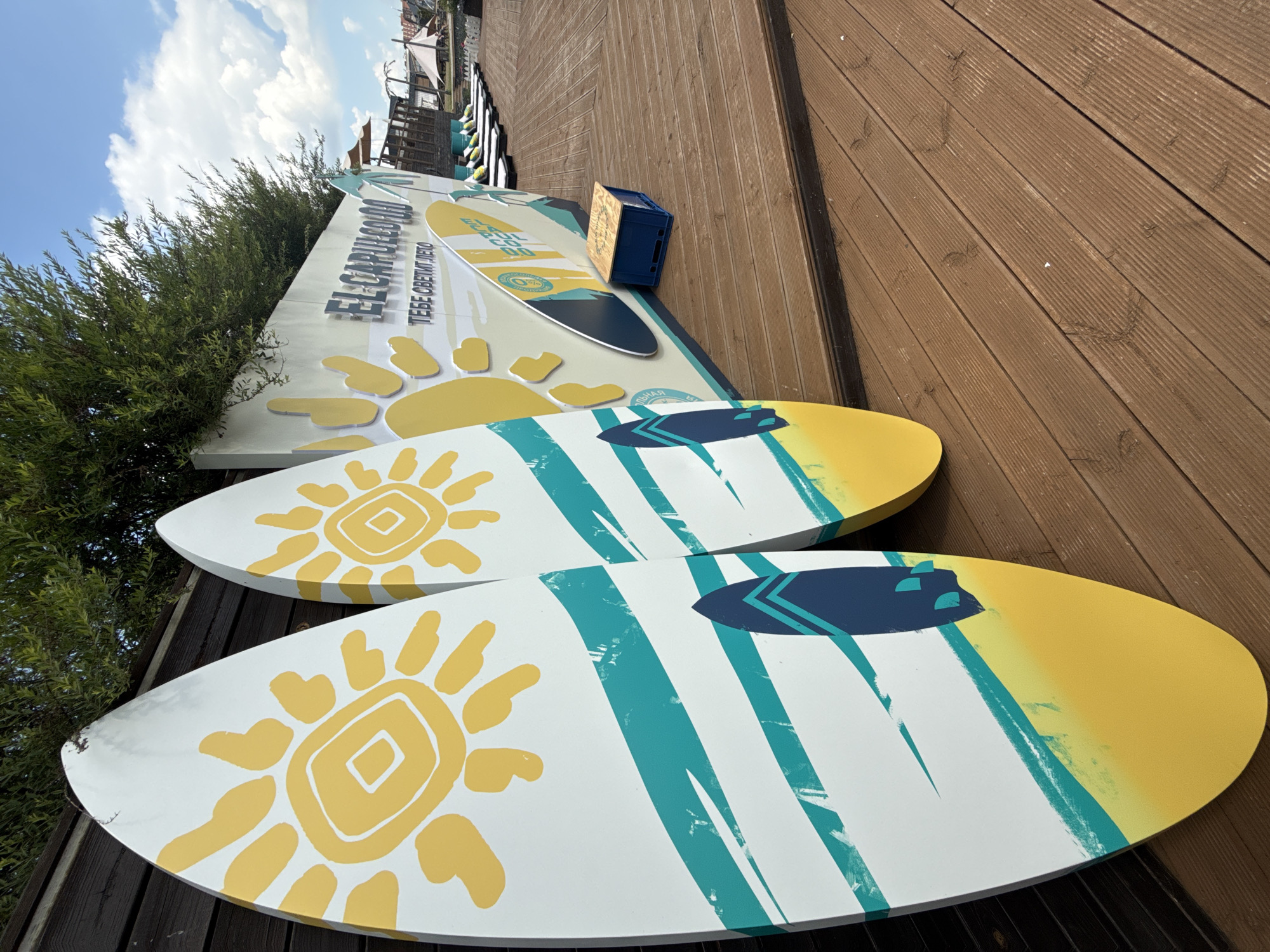 This image depicts an outdoor scene on a wooden deck, likely a boardwalk or a beach area. In the foreground, two large, decorative surfboards stand upright, leaning against a dark wooden fence. These surfboards are white with vibrant graphics: yellow suns with radiating lines, turquoise and white diagonal stripes, and a dark blue silhouette of a surfboard with turquoise accents. To the right, a light-colored wall features a large graphic with the text "EL CARPULLOOD" and "ТЕБЕ СВЕТИТ ЛЕТО" (meaning "Summer Shines For You"). Further in the background, several white lounge chairs are arranged on the wooden deck, suggesting a relaxation or recreational area. Beyond the chairs, tents and structures are visible, indicating a commercial or event space. The environment is outdoors, with lush green foliage above the wall and a blue sky with scattered clouds visible in the upper right corner. The presence of surfboards and lounge chairs suggests a beach resort, water park, or a similar leisure establishment. The text on the wall, specifically "ТЕБЕ СВЕТИТ ЛЕТО," implies a summery theme and a welcoming atmosphere. The specific location is identified as Votkinsk, Russia.