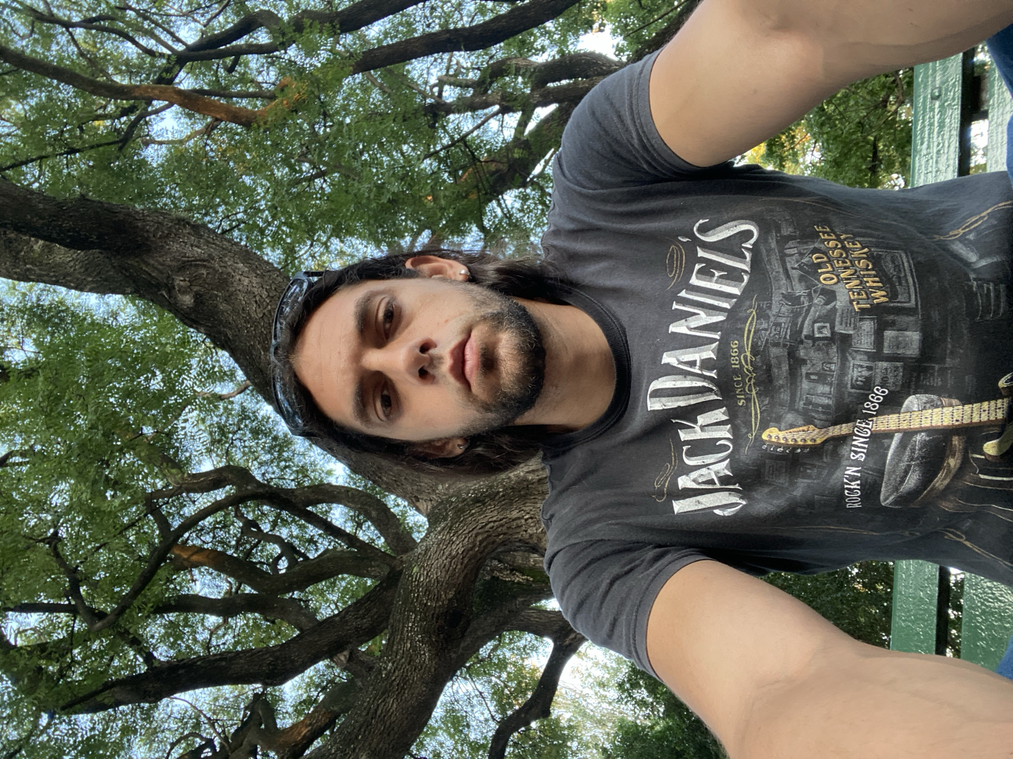 A man is taking a selfie outdoors with a large tree in the background. The man has long dark hair, a beard, and is wearing a dark grey t-shirt with a "Jack Daniel's" logo and a graphic of a guitar. The tree has thick branches with green leaves. The location cannot be determined as Buenos Aires, Argentina based on the visual information provided.