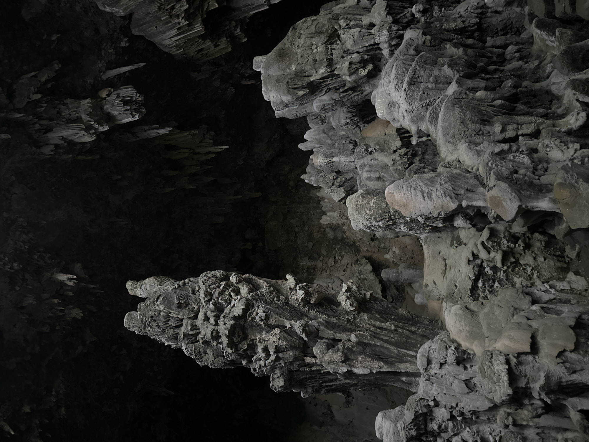 The image shows the interior of a cave in Mai Chau, Vietnam, characterized by numerous stalactites and stalagmites. The formations are predominantly grey and appear to be made of rock. The cave ceiling is dark and covered with hanging stalactites, some of which are long and thin, while others are thicker and more bulbous. On the cave floor, a variety of stalagmites rise up, with some forming large, intricate structures. The lighting is dim, casting shadows and highlighting the textures of the rock formations. There are no people or animals visible in the image. The overall environment is that of a natural cave system.