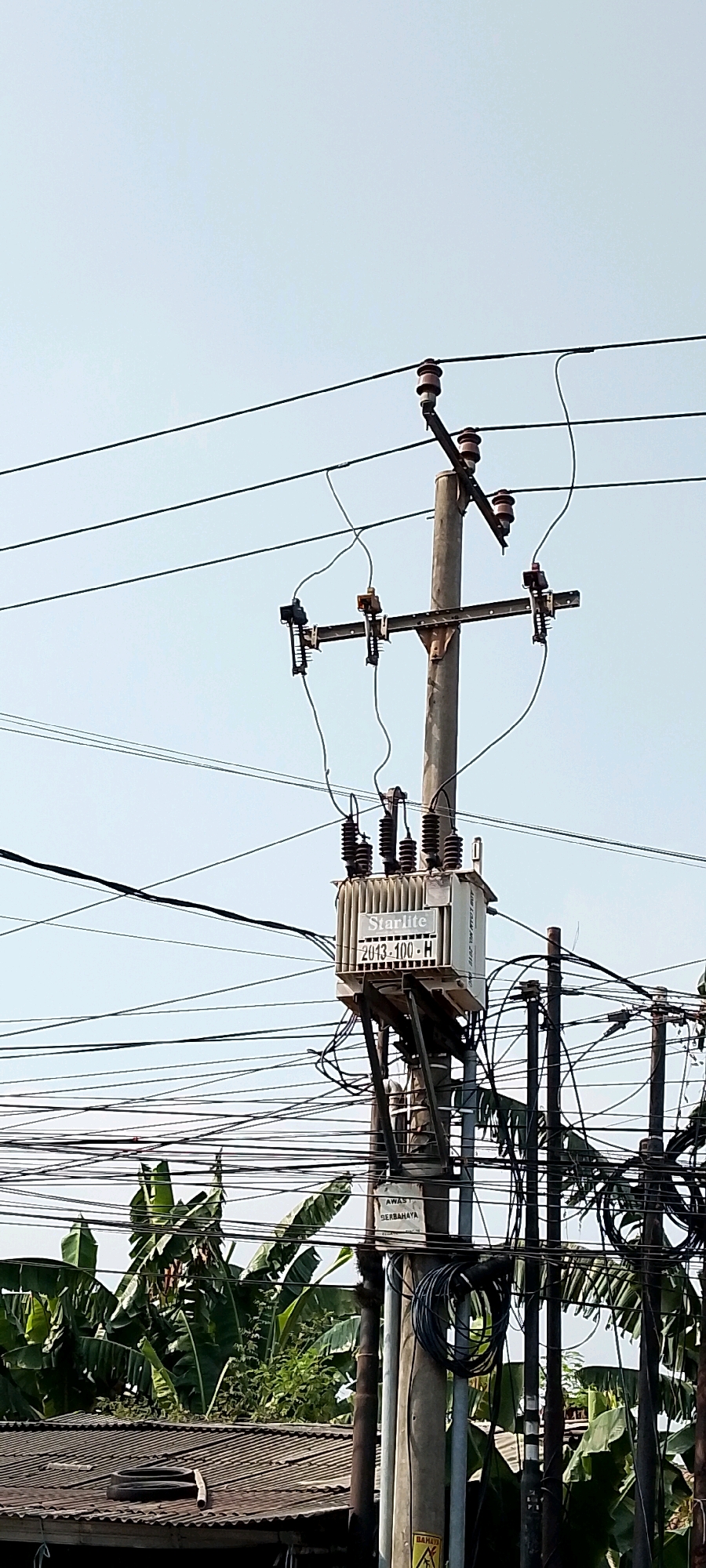 This image, taken in Karangsatria, Indonesia, depicts a utility pole with a transformer and numerous electrical wires against a clear sky. The pole itself is concrete, supporting a crossbar with ceramic insulators from which electrical wires are suspended. A beige electrical transformer unit is mounted on the pole, featuring the brand name "Starlite" and the model number "2013-100-H" on a label. Several dark, insulated wires connect to the transformer and extend to other parts of the network. In the lower portion of the image, a dense cluster of lush green banana trees and other foliage is visible, suggesting a tropical environment. Below the trees, a portion of a corrugated metal roof of a building is present. Additional utility poles and a tangled web of electrical cables fill the background, creating a dense urban infrastructure impression. A warning sign with text in a foreign language is attached to one of the poles.
