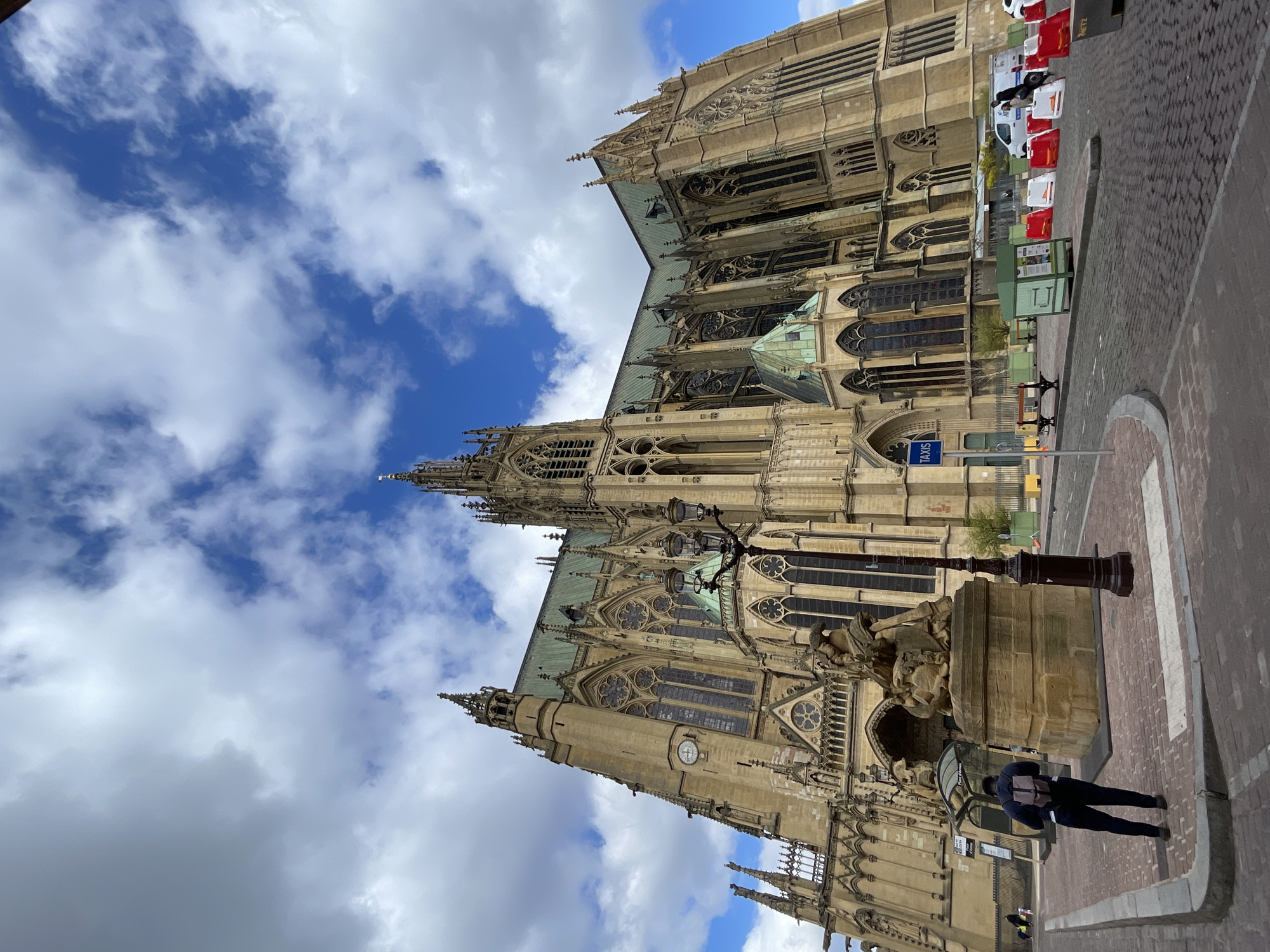 The image captures a view of Metz Cathedral, formally known as the Cathedral of Saint Stephen of Metz, located in Metz, France. The cathedral's facade, built of Jaumont stone, is prominently displayed, showcasing its Gothic architectural details, including pointed arches, stained glass windows, and ornate spires. A fountain and street lamp stand in front of the cathedral. A person is visible on the sidewalk beside the fountain, and traffic barriers line the curb further along the street. The sky features blue patches interspersed with white clouds.