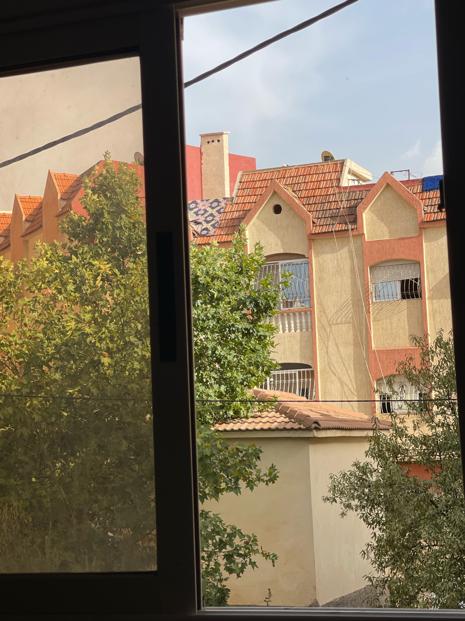 The image shows a view through a window of buildings and trees in Azrou, Morocco. In the foreground, the dark frame of the window is visible. Through the window, there is a large tree with green leaves on the left, partially obscuring a building. This building is light beige with a tiled orange roof, featuring distinctive gabled sections. Behind it, a larger building with a similar orange tiled roof is visible. This second building is painted in warm tones of yellow and coral pink, with arched windows covered by white metal bars. Further in the background, a tall, light-colored chimney rises from a red-tiled roof. A satellite dish is visible on the roof of a building to the right. The sky is light blue with some scattered clouds. The overall scene depicts an urban residential area with a blend of architectural styles and lush greenery.