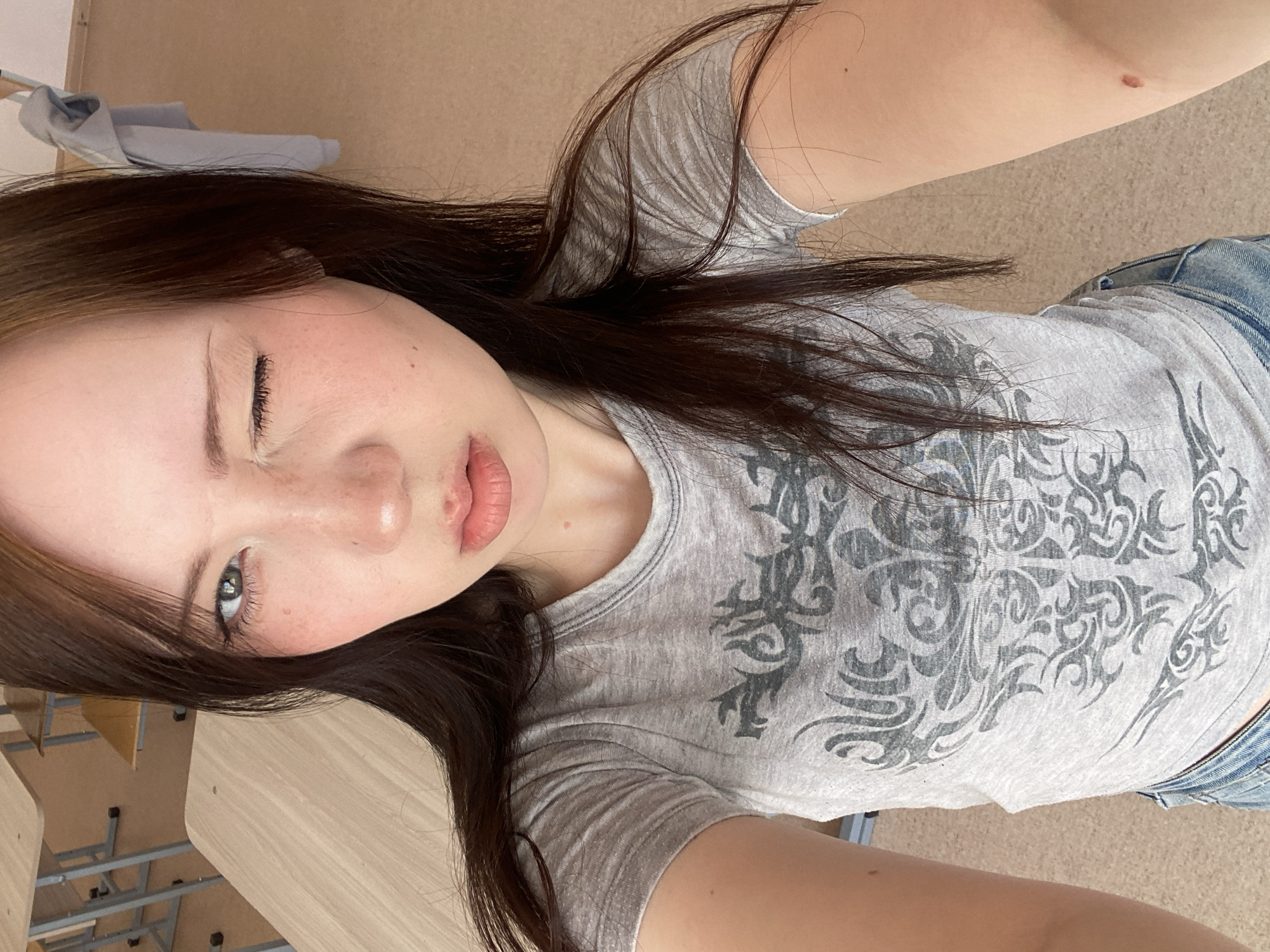A selfie taken by a young woman with fair skin and brown hair. She is wearing a gray t-shirt with a patterned design and blue denim jeans. The woman is winking with her left eye closed. The background shows a classroom with tables and chairs. The location is Unknown, Unknown.