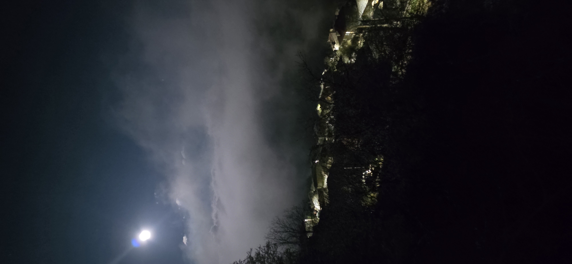 The image shows a nighttime scene in Breze, Bulgaria. In the foreground, there is a dark area with silhouettes of trees or foliage. To the left, the sky is visible with dark clouds and a bright moon. In the middle ground, there is an area of fog or mist. Beyond the fog, there are lights visible, suggesting the presence of buildings or structures. The image appears to be taken at an angle, likely from a lower vantage point looking upwards towards the town.