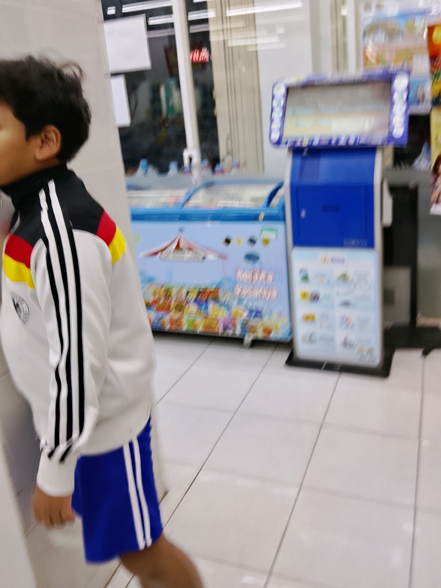 The image captures a young boy wearing a white and black jacket with red and yellow accents on the shoulders, paired with blue shorts. He is standing in what appears to be a convenience store, indicated by the tiled flooring and the presence of a freezer unit and a modern payment kiosk. The freezer is adorned with colorful graphics, possibly advertising ice cream or other frozen treats, featuring a circus tent motif. To the right, a blue payment kiosk displays information on a screen, with promotional posters nearby.
The boy is partially visible, with his head and upper body in the frame, and he is facing away from the camera, looking towards the freezer and kiosk. The scene is indoors, under artificial lighting, suggesting it could be any time of day. The overall impression is a casual, everyday shopping experience. There is no visible text that can be clearly deciphered from the image. The location context of Malang, Indonesia, suggests this is likely a store within that city.