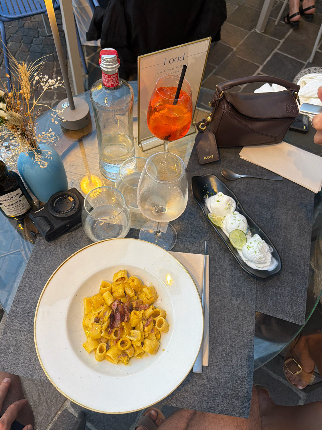 The overhead shot depicts a dining scene on an outdoor patio in Loveno, Italy, likely at dusk or during evening hours, with ambient lighting. In the foreground, a white ceramic plate with a golden rim holds a dish of pasta with a creamy yellow sauce and pieces of bacon. To the right, a black oblong dish contains two scoops of white ice cream garnished with lime slices and zest. A silver spoon rests beside the ice cream.
The table is covered with a dark gray textured placemat. Several wine glasses, some containing white wine, are scattered around the table. A bottle of clear liquid, possibly spirits, stands upright. Adjacent to the bottle is a table lamp with a concrete base. A small bouquet of dried flowers and wheat in a light blue vase sits to the left of the lamp. A black compact camera is placed on the table near the flowers.
A dark brown leather handbag, featuring the Loewe logo on a detached tag, is positioned to the right of the bottle and in front of a small stand displaying the word "Food." An orange cocktail with ice and a black straw sits in a stemmed glass. A folded white napkin and a silver utensil are also visible.
The lower half of the image shows the legs and feet of at least two individuals, suggesting they are seated at the table. One person's legs are visible in the bottom left, wearing dark shorts. Another person's legs and feet, wearing sandals with gold buckles, are visible in the bottom right. The background is blurred, hinting at a European-style streetscape with cobblestones and building facades.