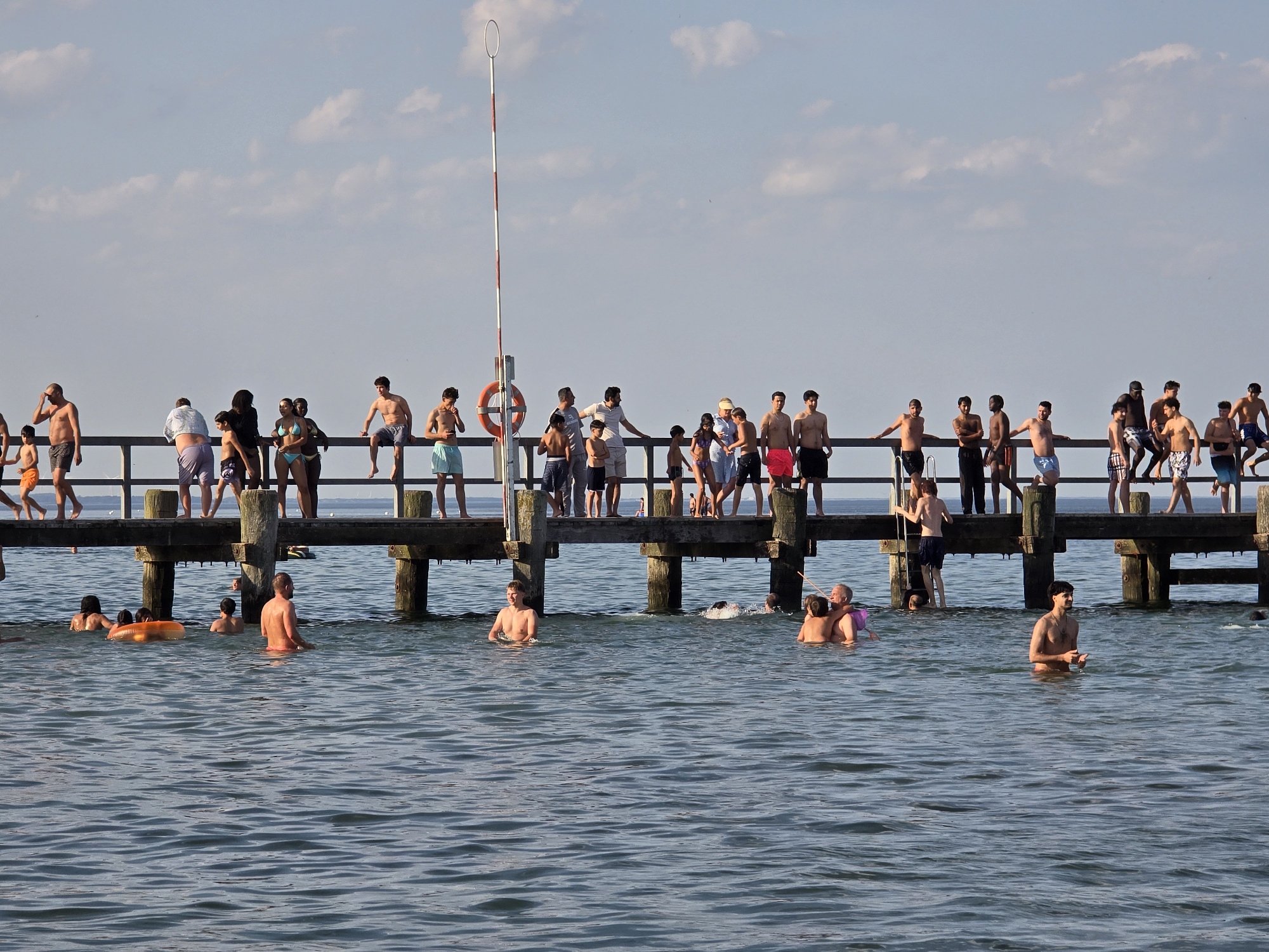 In the city of Lübeck, Germany, on the Baltic Sea coast, a wooden pier extends over the water. A diverse group of people, primarily in swimwear, are seen both on the pier and in the water. On the pier, individuals are standing, sitting, and leaning against the railing, observing the scene below or interacting with each other. Some are dressed in casual beach attire, while others are shirtless. In the water, people are swimming, wading, and floating, with some engaged in playful activities. There is an orange inflatable ring visible in the water. The sky above is a light blue with scattered white clouds, indicating fair weather. The overall atmosphere suggests a leisurely summer day at the beach.