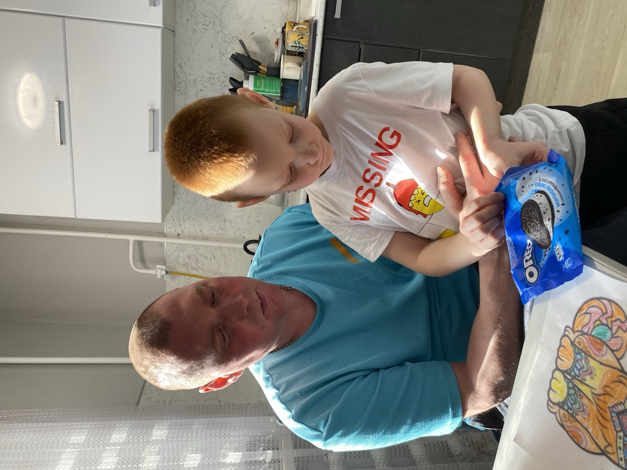In an indoor scene, a man and a young boy are closely positioned. The boy, with red hair, is wearing a white t-shirt adorned with the word "MISSING" and a cartoon graphic. He holds a package of Oreo cookies in his hands. The man, in a blue shirt, is seated and appears to be supporting the boy. In the foreground, a colored drawing is visible. The setting appears to be a home environment, with kitchen cabinets visible in the background. The location is Noviy, Russia.