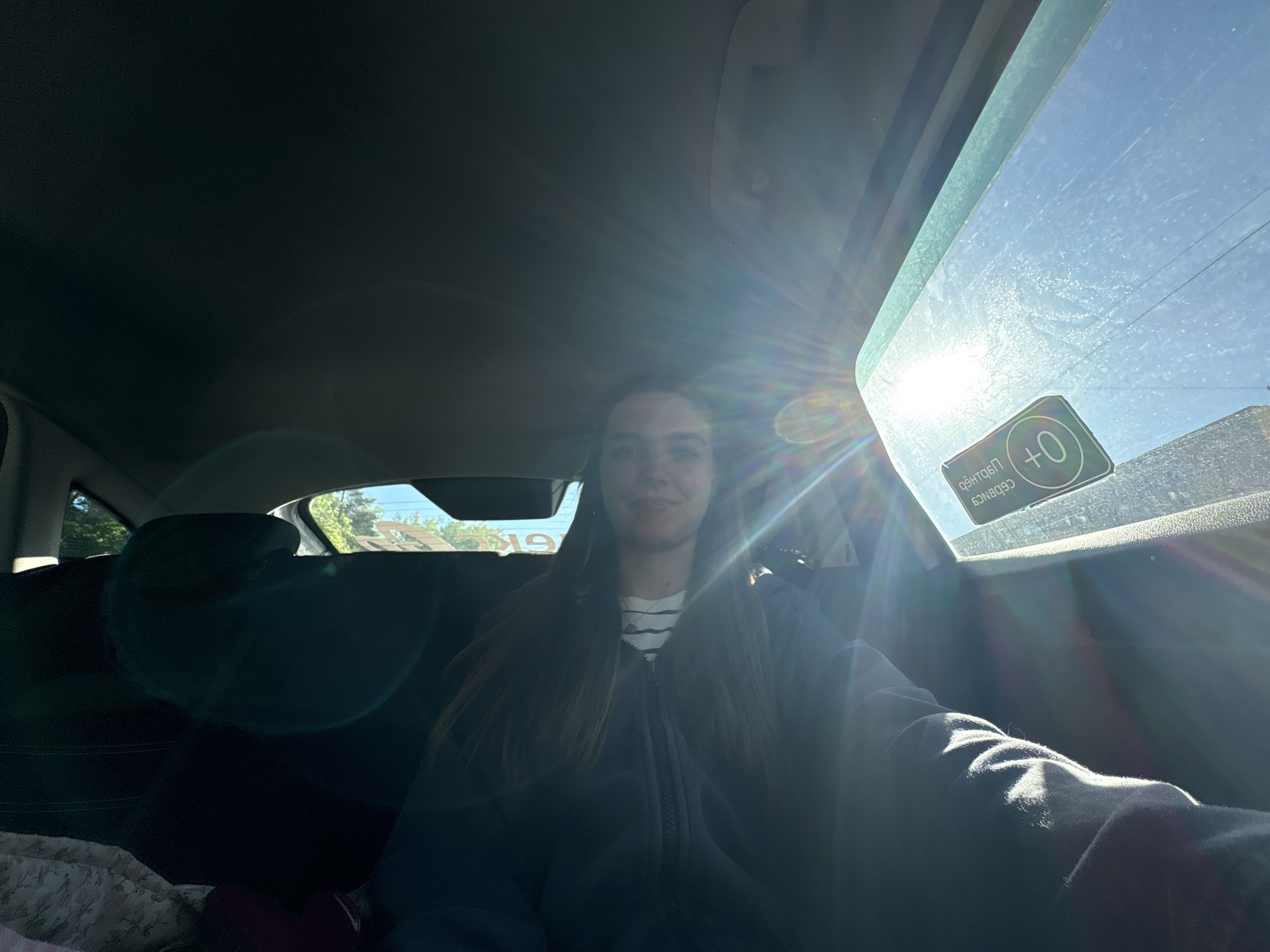 A young woman is seated in the driver's seat of a car. She is wearing a striped shirt and a dark-colored zip-up hoodie. The bright sunlight streams through the front passenger window, obscuring the scene. A sticker with "+0" is visible on the window. Based on the presence of the sticker, the car is likely located in Perm, Russia. The back seats are partially visible. The image appears to be taken from the driver's point of view.