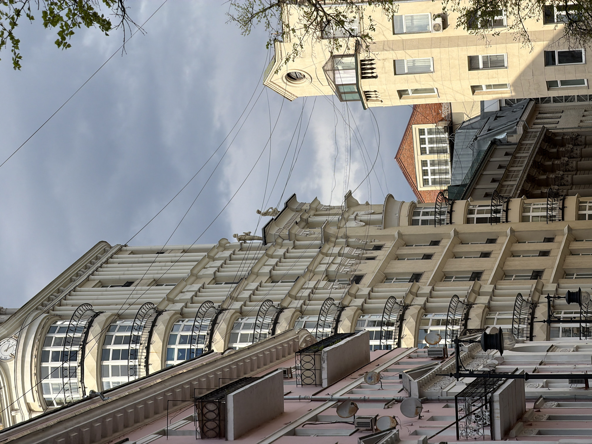 The image, taken from a low angle, showcases the architecture in Moscow, Russia. The facades of several buildings dominate the view. One building, beige in color, features a repeating pattern of arched windows with decorative black metal railings. Above the windows are numerous angled ledges. Further up, the building is adorned with sculptures. To the right of this building, a portion of a cream-colored building with a bay window and standard rectangular windows is visible. A smaller red-brick building with a gabled roof is nestled in between the two taller structures. Overhead, several power lines crisscross the sky, which is overcast with grey clouds. Bare tree branches with sparse green foliage are visible in the top left and right corners of the frame. The image suggests a mix of architectural styles within a densely populated urban environment.