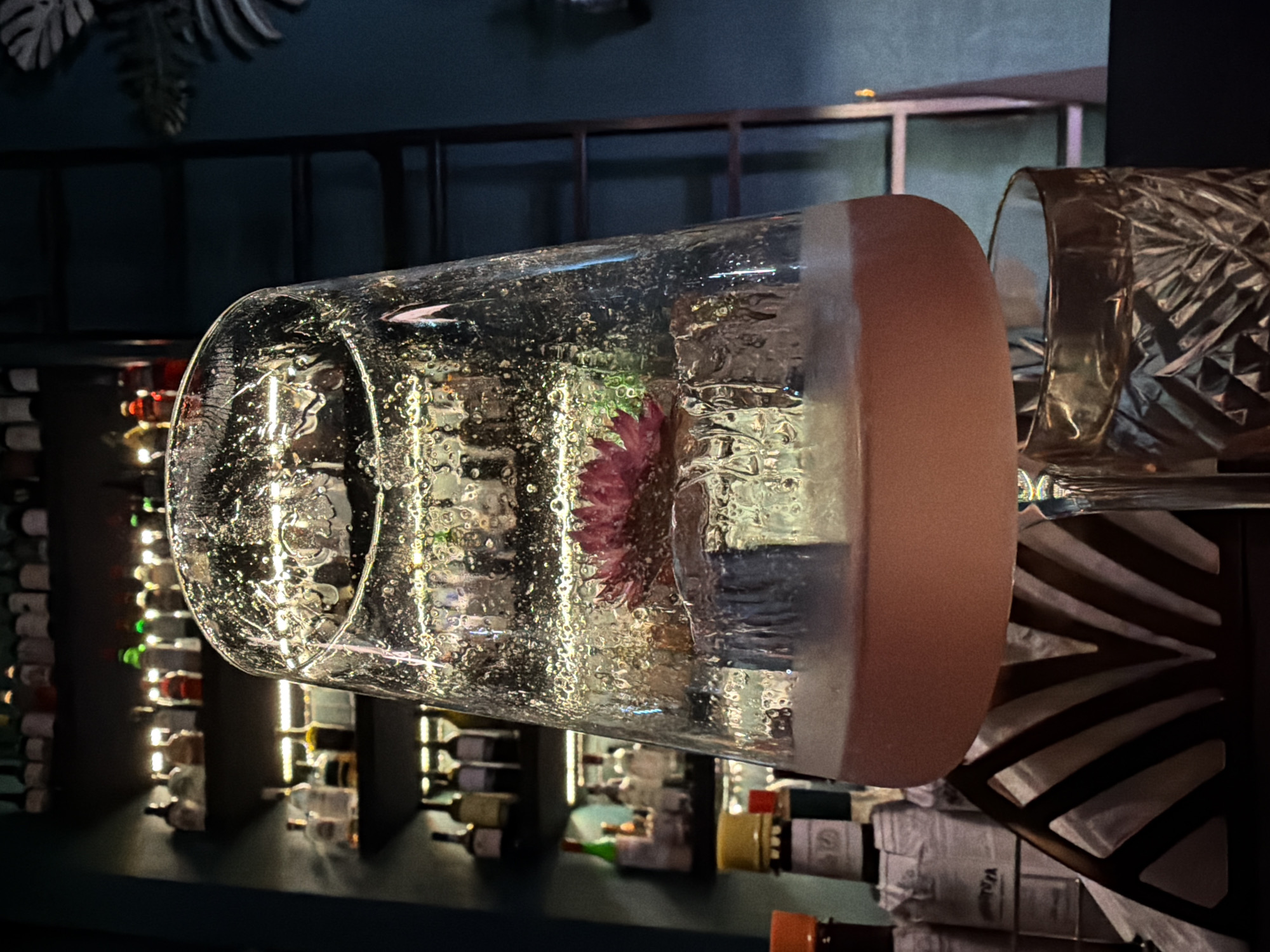 A close-up, horizontally oriented shot captures a unique drink presentation in a dimly lit bar or restaurant setting. The central focus is a tall, clear glass, possibly a highball, filled with a beverage and a large amount of textured ice. A vibrant red or magenta flower, along with a small green leaf, is artfully encased within the bubbly, irregular ice. The rim of this glass features a frosted or opaque band in a muted orange-pink color.
To the immediate right of the main glass, a portion of another glass is visible, characterized by a distinct cut-glass or etched pattern, possibly an old-fashioned glass. The background on the left reveals shelves densely packed with numerous bottles, likely wine or spirits, which are illuminated from behind, casting a warm glow. The upper background on the right is dark, showing hints of structural elements like metal bars and what appear to be decorative, dark-colored leaves. The overall lighting is low and atmospheric, highlighting the intricate details of the cocktail's presentation. Text on bottle labels in the background is present but indistinguishable.