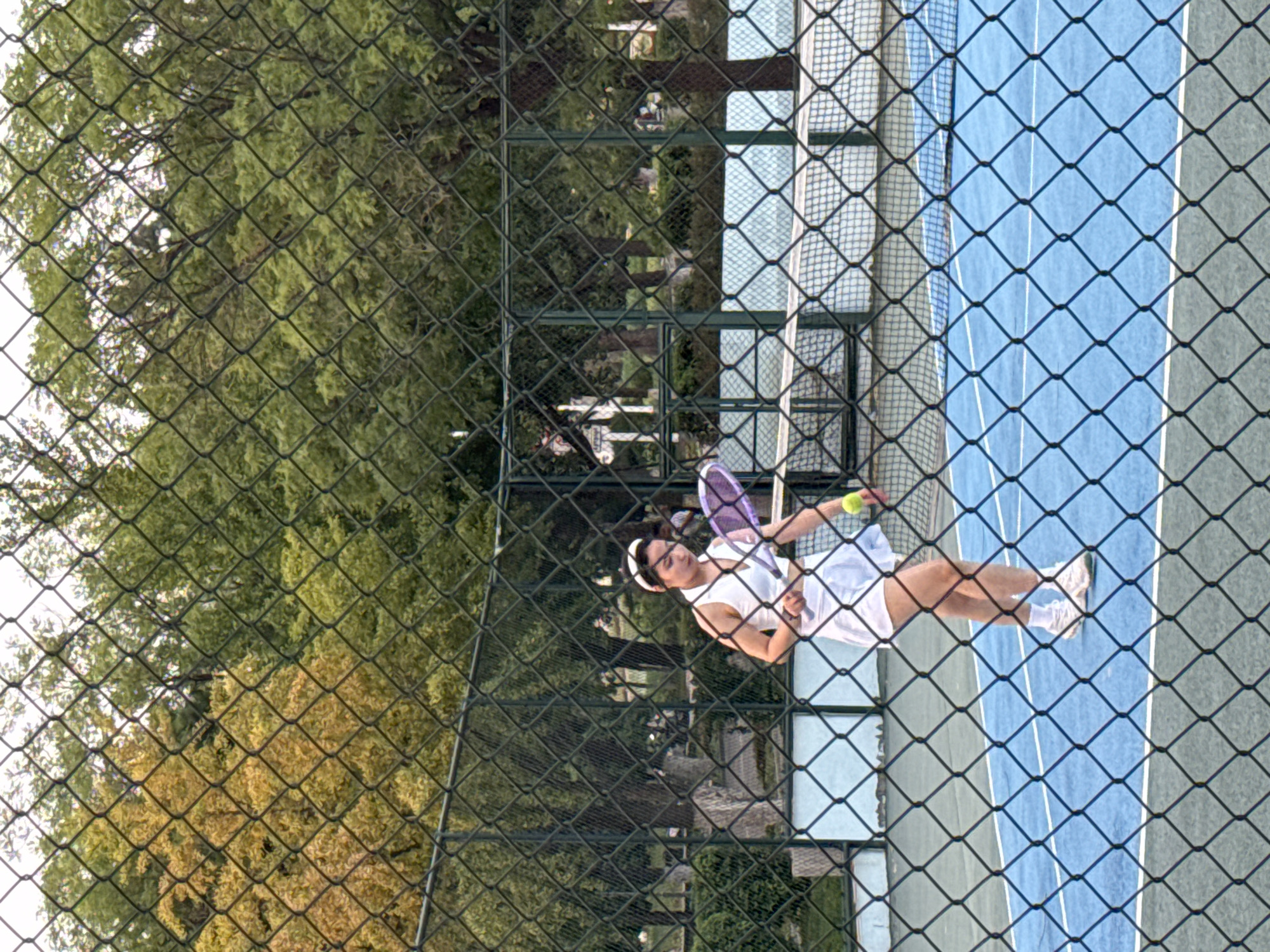 A young woman is captured mid-play on a blue tennis court, viewed through a chain-link fence. She wears a white tennis outfit and a white headband, and holds a purple tennis racket. A yellow tennis ball is in the air near her. The court is outdoors, surrounded by trees, some of which have yellowing leaves, suggesting it may be autumn. The setting appears to be a public park or recreational area in Linghe, China.