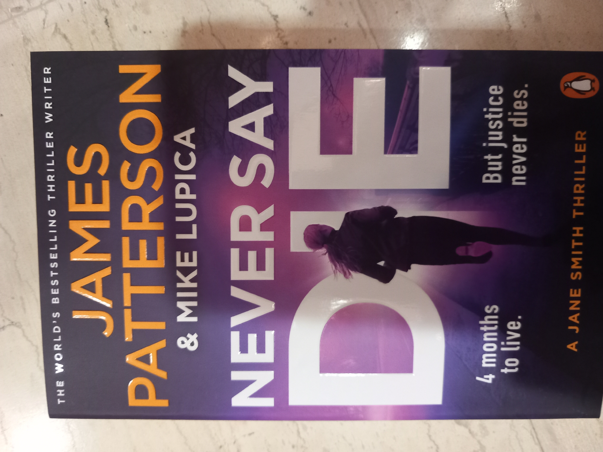 A book cover for "Never Say Die" by James Patterson and Mike Lupica is displayed. The cover is primarily dark purple with vibrant purple and pink accents in the background. Large, bold white lettering dominates the cover, spelling out "NEVER SAY DIE." A silhouette of a woman running is positioned in the center of the title. Below the title, in smaller white text, it reads "4 months to live." To the right, also in white text, is "But justice never dies." At the bottom of the cover, in orange text, it says "A JANE SMITH THRILLER," with a Penguin Books logo to its right. The book cover is lying flat on a surface that appears to be a table or floor with a subtle marble-like pattern. There are no people, actions, or interactions depicted outside of the book cover's design. The environment is an indoor setting.