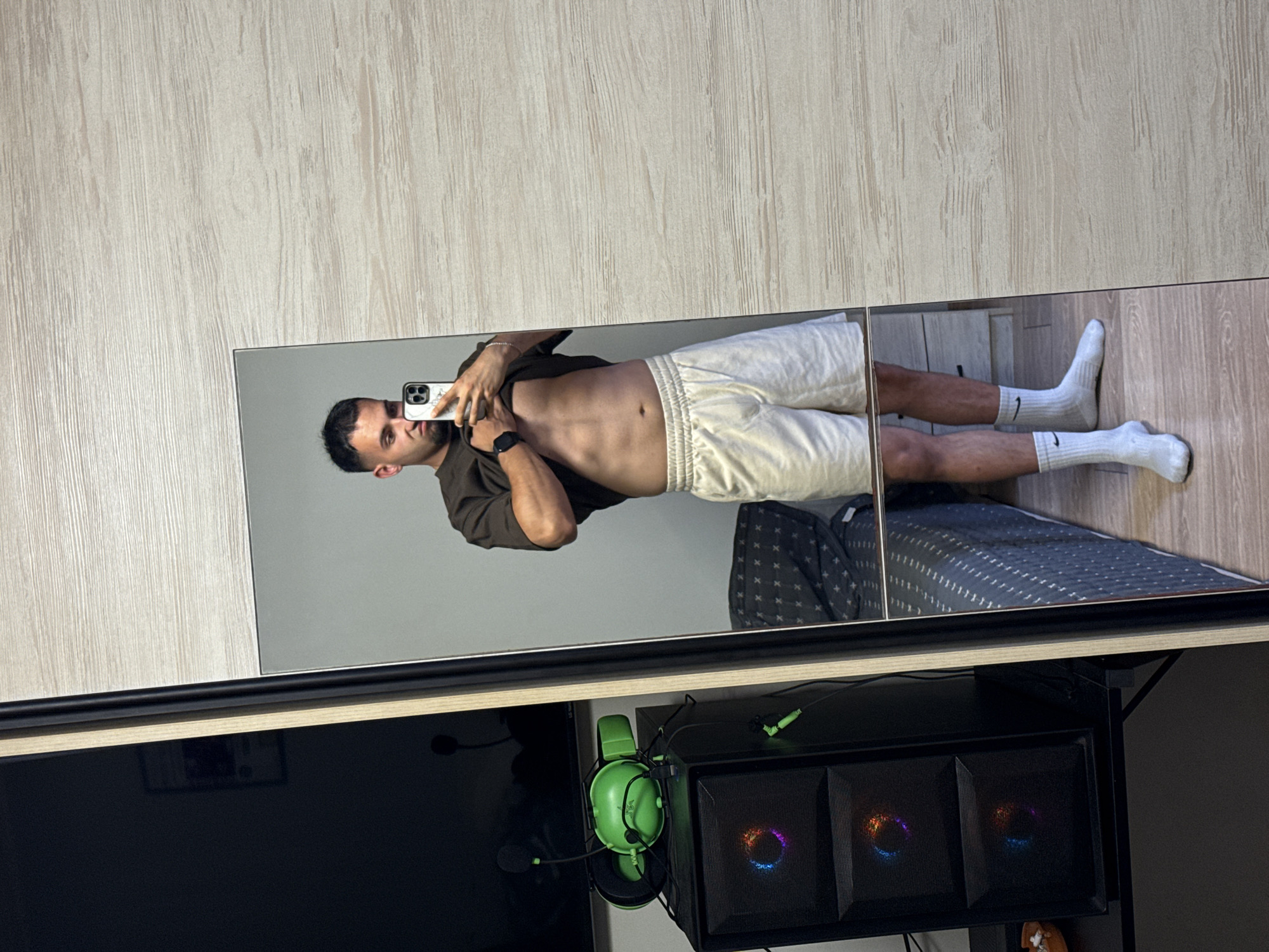 The media file displays a mirrored reflection of a man in athletic attire, standing in what appears to be a bedroom or living space. The man is wearing a dark t-shirt, light-colored shorts, and white socks. His physique suggests he is fit, with visible abdominal muscles. He is holding a smartphone in his right hand, angled to capture the reflection. A watch is visible on his left wrist.
The setting includes a light-colored wooden floor and a wall with a wood-grain texture. Below the mirror, a black computer tower with illuminated fans and a black monitor are partially visible, suggesting a home office or entertainment area. A pair of bright green headphones with a microphone is hanging from the side of the computer tower. The lighting suggests it is daytime, possibly indoors. No specific weather conditions are indicated. The overall mood conveyed is casual and personal, as the man is taking a self-portrait. There is no visible text in the media file. The location context provided is Shumen, Bulgaria.